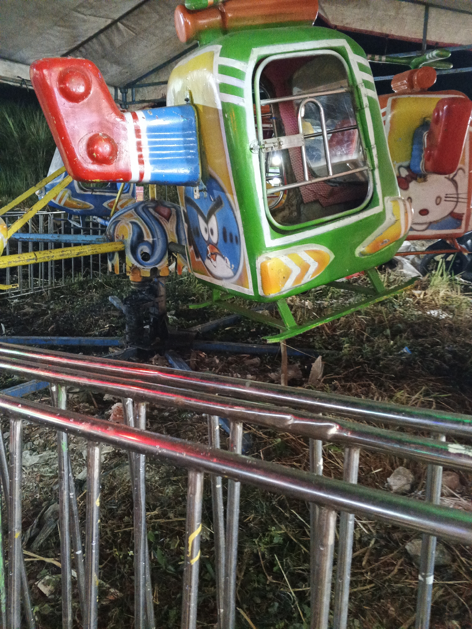 A close-up, low-angle shot captures a section of a children's amusement ride, likely a carousel with helicopter-shaped pods, located outdoors in Sukabumi, Indonesia. The ride features a green and white helicopter pod with orange accents and a cartoonish blue bird painted on its side. The pod has a transparent window with a metal safety bar. To the right, another pod is partially visible, adorned with a Hello Kitty decal. The base of the ride's central mechanism is visible, with exposed wires and metal components. In the foreground, a chrome-plated metal railing, likely a safety barrier for the attraction, is prominent, reflecting red light. The ground beneath the ride is covered in dry grass, dirt, and debris, suggesting an outdoor setting that may not be meticulously maintained. The overall lighting suggests it is either dusk or night, with artificial lights from the attraction casting a glow.
