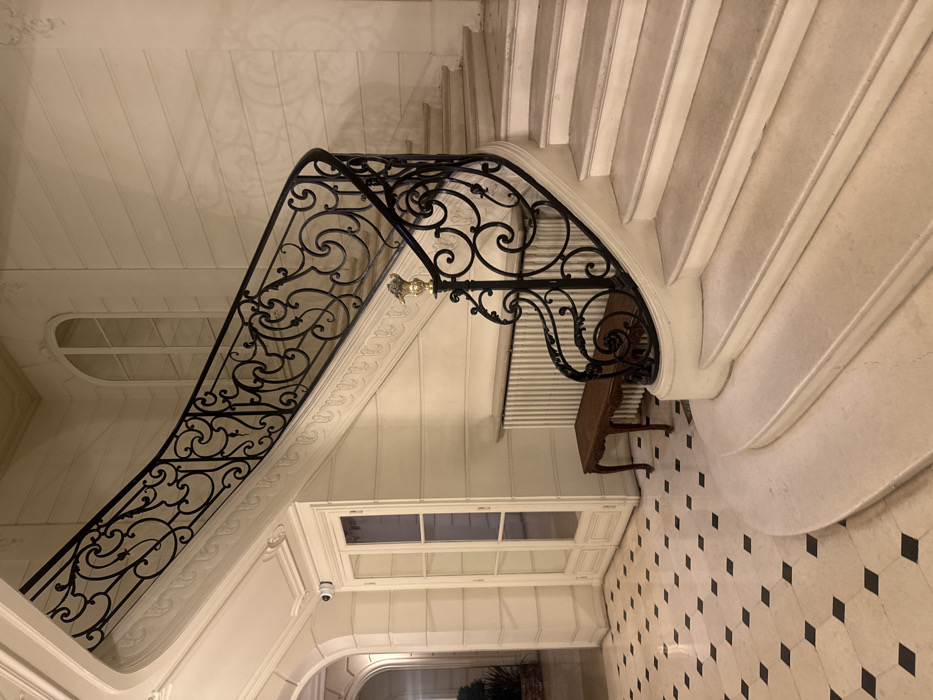 The image depicts a grand interior staircase within what appears to be a historic, elegant building in Paris, France, likely a private residence, embassy, or institution. The scene is devoid of individuals.
A curved staircase with light-colored, possibly marble or stone, steps ascends from the lower right of the frame towards the upper right. It features an ornate black wrought-iron balustrade with intricate scrollwork and floral motifs. A brass or gold-colored finial, detailed with a small figure, crowns the newel post at the bottom of the balustrade.
The walls are painted in a light cream or off-white color, featuring horizontal paneling and classical moldings. To the left of the staircase, a tall, white double-door with multiple glass panes is visible, above which a small white security camera is mounted. Below the rising stairs, a long, white, fluted radiator is installed against the wall, with a small, dark-wood decorative bench positioned in front of it. Further left, a large arched passage leads to another illuminated interior space, where a potted plant is partially visible. An arched window with multiple panes is visible on the upper wall, suggesting a light source. The floor is covered with large, light-colored tiles interspersed with smaller black square tiles arranged in a diamond pattern.