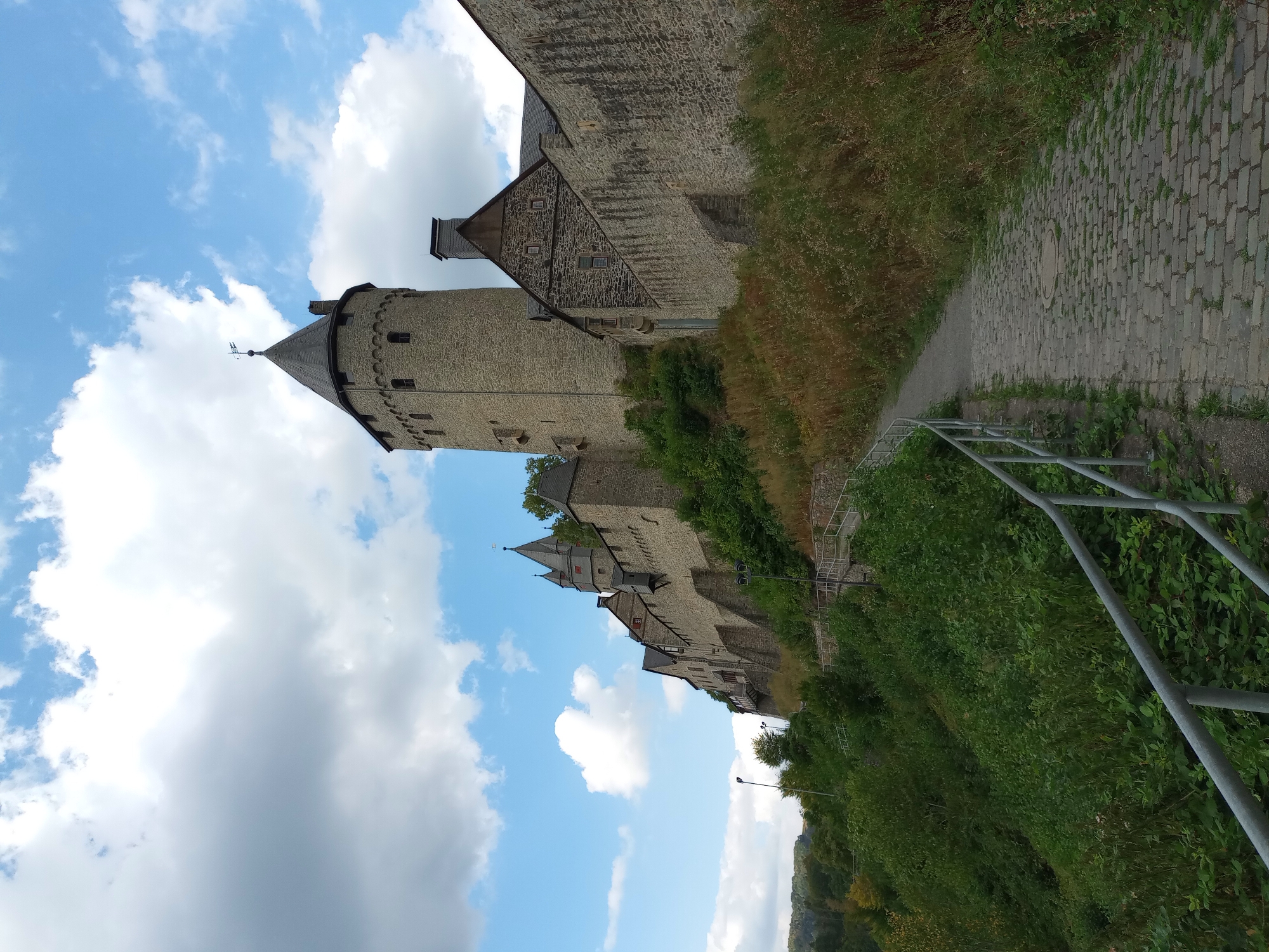The image shows a view of Altena Castle in Altena, Germany, from a cobblestone path leading up to it. The castle is perched on a hilltop, with a prominent stone tower and crenellated walls dominating the skyline. The architecture is medieval, with stone construction and pointed roofs. The path is lined with a metal railing and bordered by lush green vegetation and tall, dry grasses. The sky above is blue with scattered white clouds. There are no people visible in the image. The overall impression is one of historical grandeur and natural beauty.