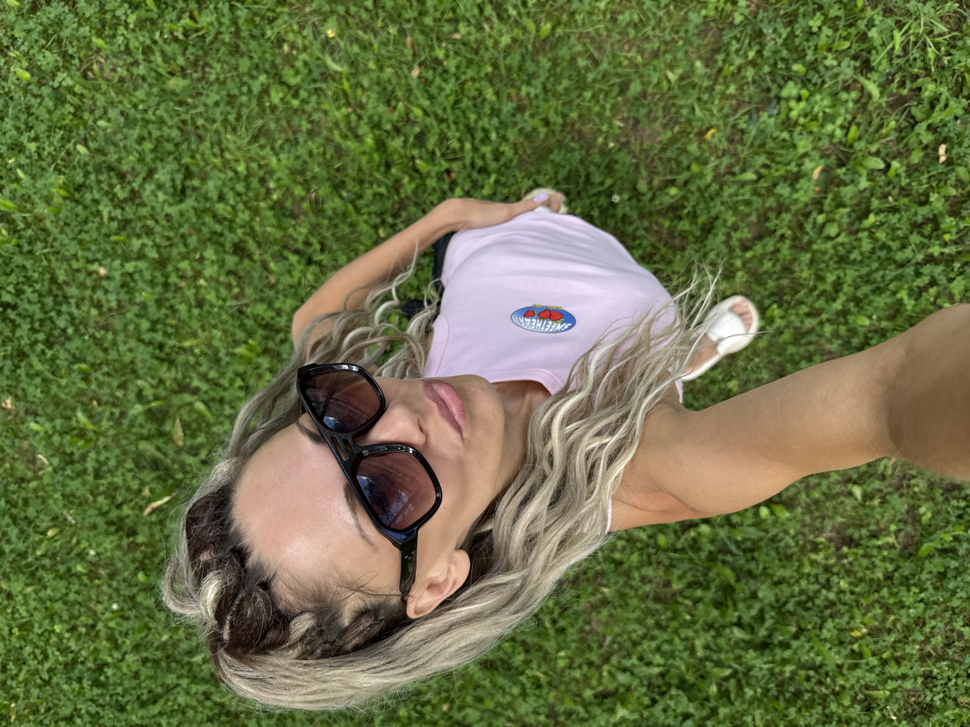 An overhead shot captures a woman posing for a selfie on a grassy lawn. She is positioned in the center of the frame, looking up and slightly to the left. The woman has long, blonde, braided hair pulled back into a ponytail, with some strands framing her face. She is wearing large, dark-rimmed sunglasses and a light pink tank top with a circular logo featuring red strawberries and the text "SWEETHEART". She has on white sandals. Her right hand is extended to hold the camera. The background is a dense green lawn, suggesting an outdoor setting, possibly in a park or yard. The lighting indicates it is daytime, with shadows visible on the grass. No other individuals are present in the scene. The overall mood is casual and relaxed.