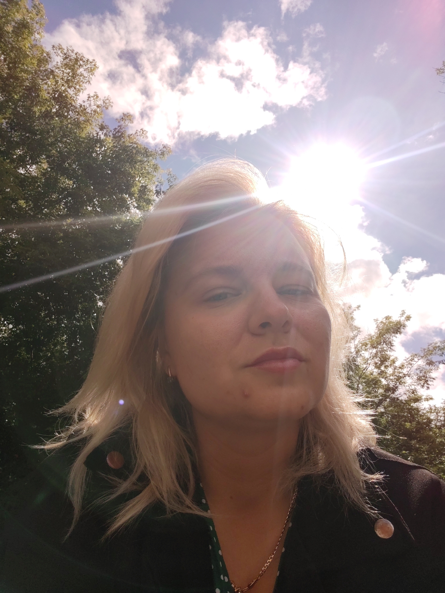 In Izhevsk, Russia, a woman with blonde hair and fair skin poses for a selfie outdoors. She is looking directly at the camera with a neutral expression. The sun is shining brightly from the upper right portion of the frame, creating lens flare and illuminating her face and hair. She is wearing a dark jacket over a green dress with white polka dots. A gold chain necklace is visible around her neck. Behind her, green trees and a blue sky with white clouds form the backdrop.