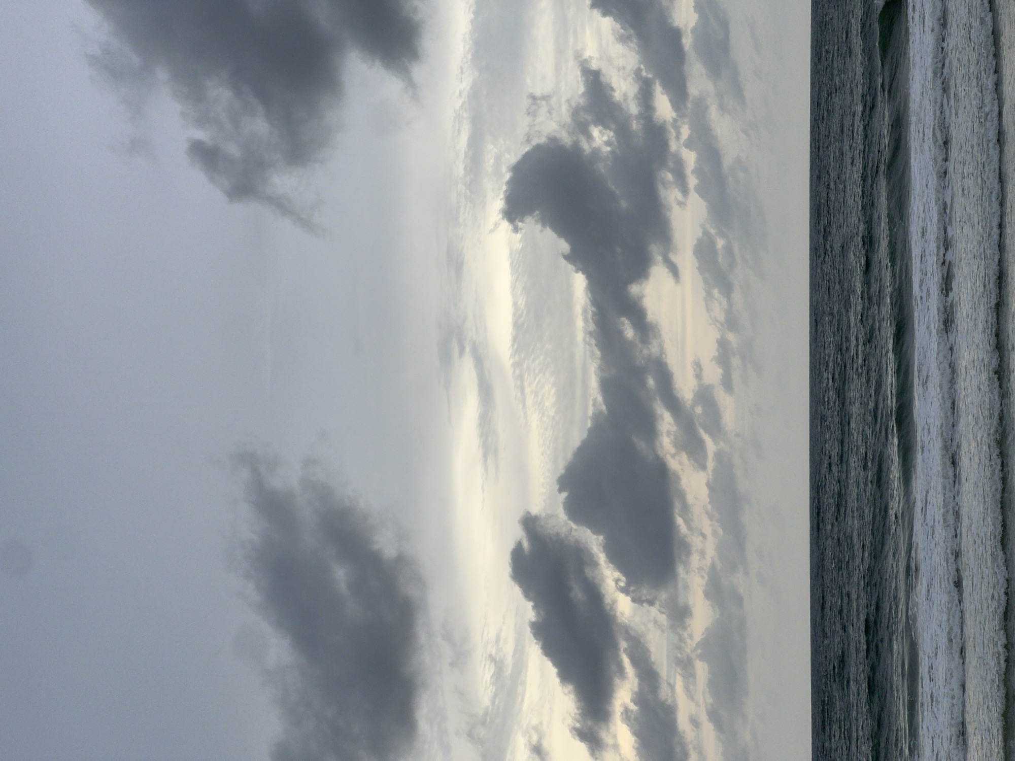The image depicts a coastal scene with a cloudy sky and ocean waves. The sky occupies the majority of the image, featuring a variety of grey clouds against a lighter, overcast background. To the right of the frame, the ocean is visible, with waves creating a textured pattern in varying shades of grey and white. The image appears to be taken on a cloudy day, with a muted color palette overall. The location is unknown.