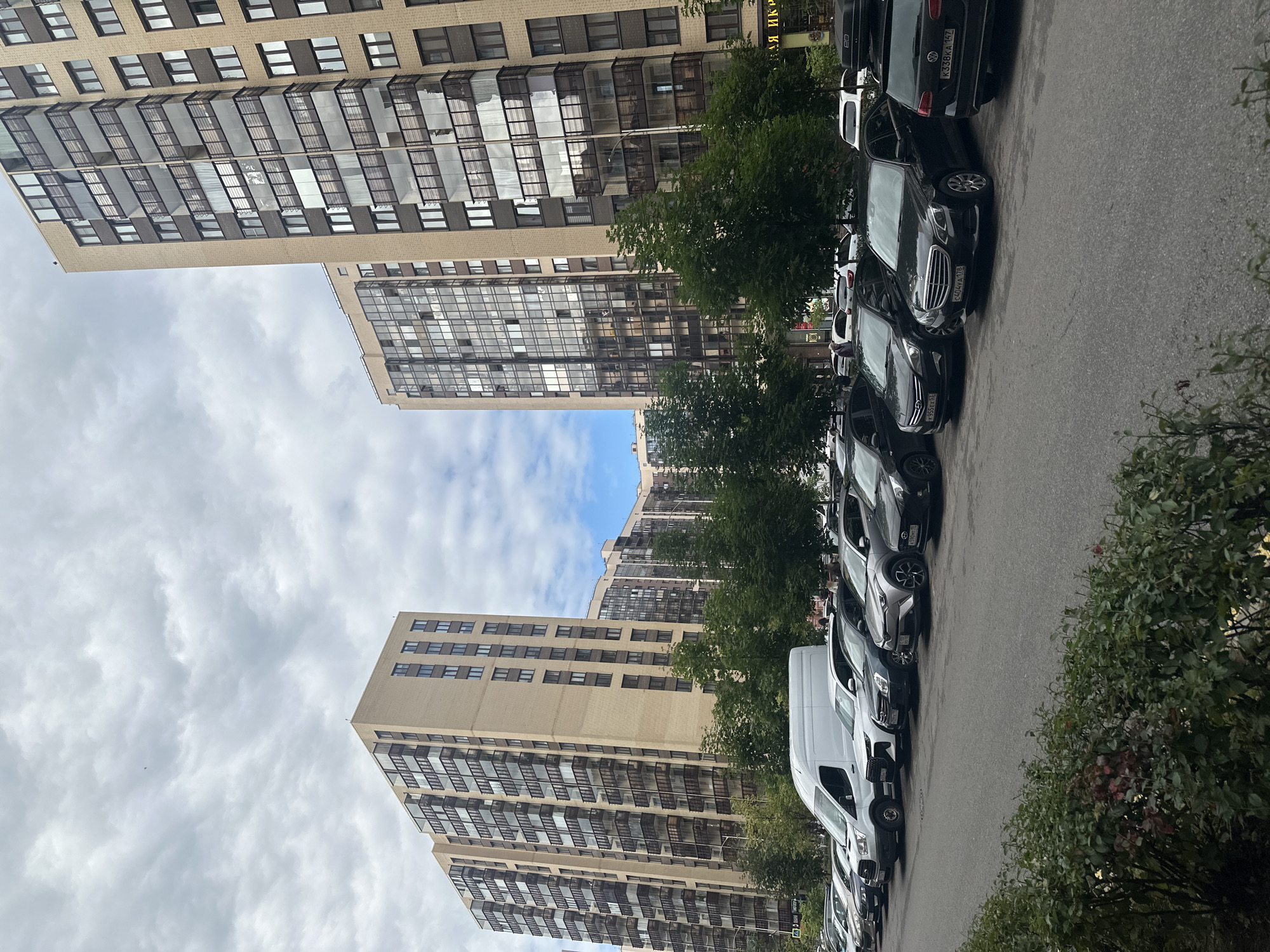 This image captures a street scene in Murino, Russia, featuring several modern apartment buildings and a row of parked cars. The buildings are tall, light-colored structures with multiple floors characterized by numerous windows and enclosed balconies. Trees line the sidewalk in front of the buildings, and the sky is overcast with a mix of clouds.
In the foreground, a variety of cars are parked parallel to the street. These include a white van, several sedans and SUVs in various shades of gray and black. Some of the vehicles have visible license plates. A dense green hedge runs along the bottom edge of the frame, partially obscuring the view of the lower parts of the parked cars. The overall impression is of a residential area with contemporary architecture and everyday urban activity.