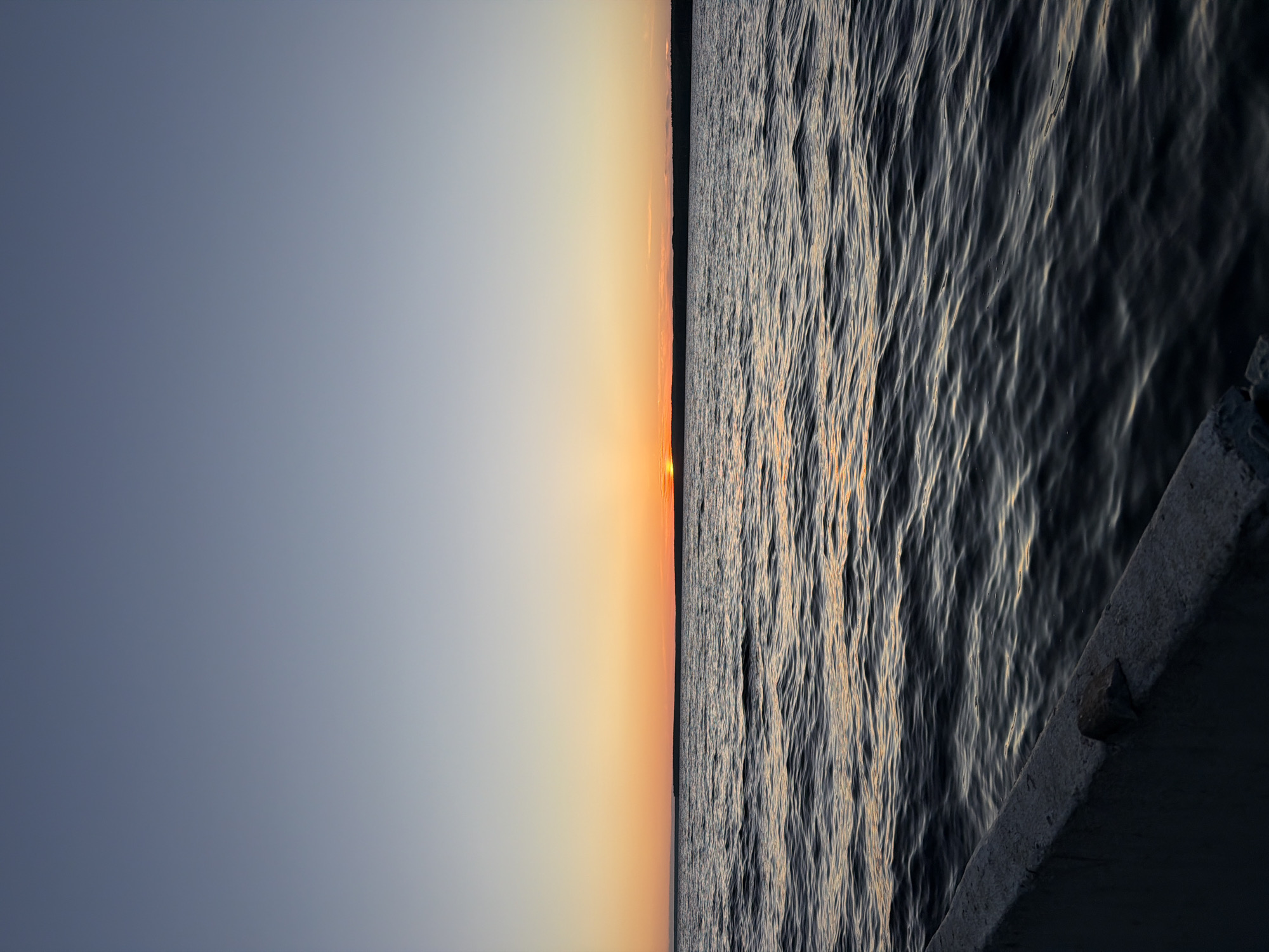 This is a vertical, eye-level, medium shot of a body of water with a sunset in the background. The water is dark and rippled, reflecting the colors of the sky. In the foreground, a concrete edge of what appears to be a pier or embankment is visible. The sky is a gradient of colors, starting with a pale blue at the top, transitioning to orange and yellow near the horizon, and finally a deep orange where the sun is setting. The sun itself is a small, bright orange disk just above the horizon line. A dark, tree-lined landmass is visible in the distance, just above the water. The scene appears to be taking place in Chaikovsky, Russia, likely on the Kama River during sunset. There are no people or animals visible in the image.