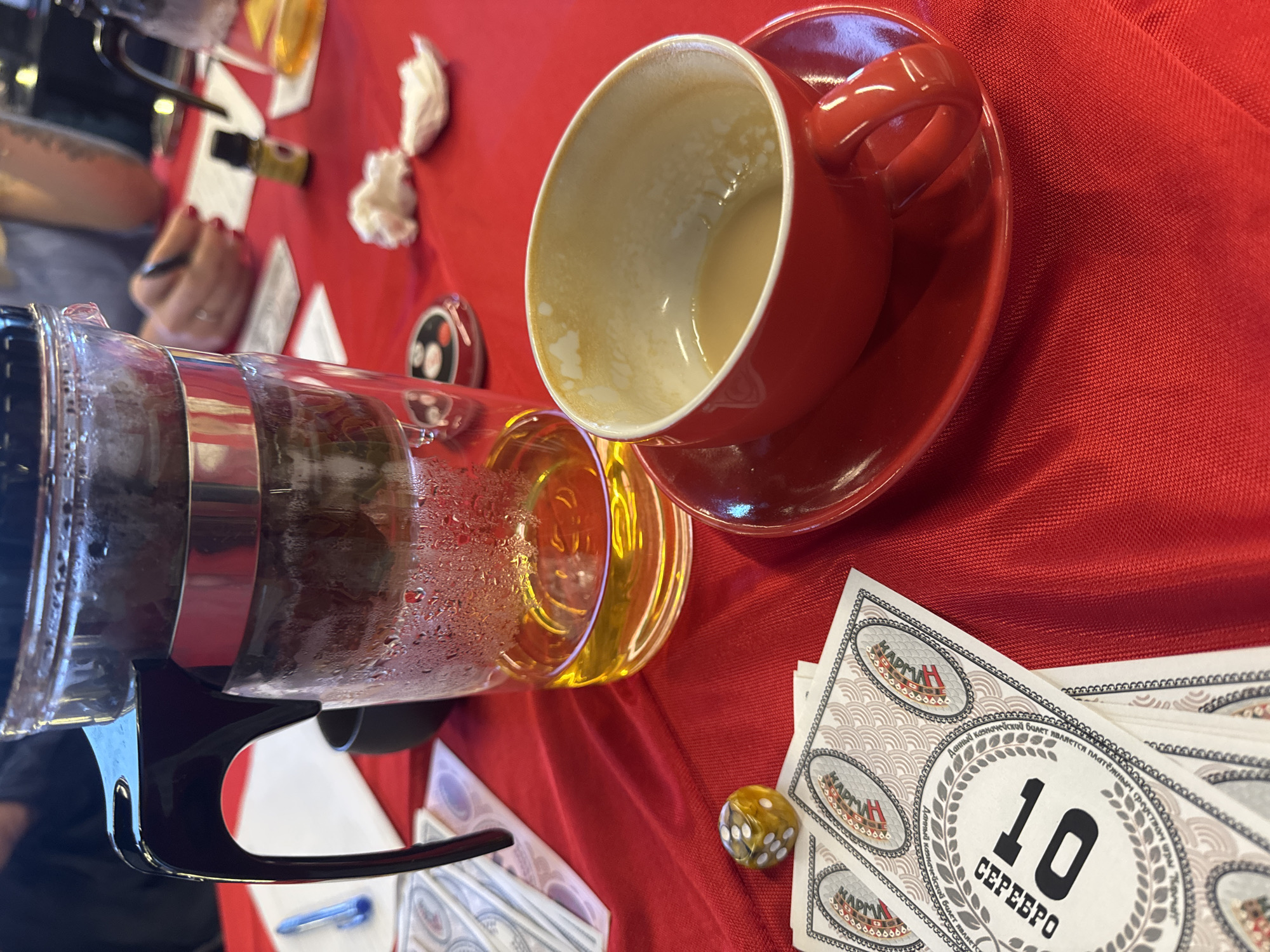 A red-clothed table in Kuta Selatan, Indonesia, holds a clear glass French press, a glass of amber liquid, and a red coffee cup with a saucer containing the remnants of a milky beverage. Beside them are stacks of paper money marked "10 CEPEEPO" and a yellow die. A person with red fingernails is partially visible in the background, holding a pen over a white document. A small bottle with a black cap and other indiscernible objects are scattered on the table.