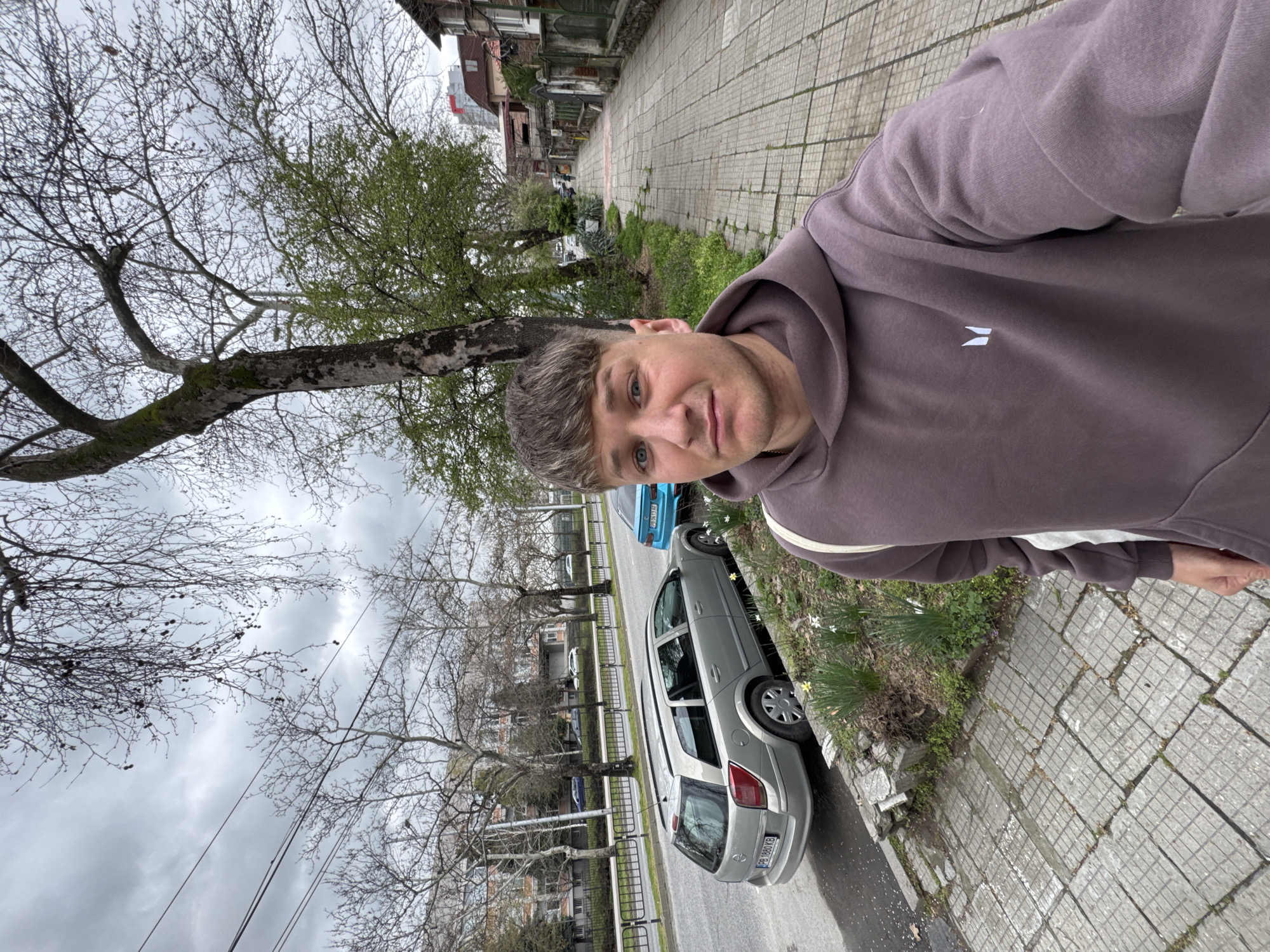 A man is taking a selfie on a sidewalk. The sidewalk is paved with square tiles and runs alongside a street. A silver hatchback car is parked on the side of the road, partially on the sidewalk. A second blue car is also parked on the street. Trees with bare branches line the street, some beginning to sprout new leaves. Buildings are visible in the background. It is overcast. The car's license plate reads "PB 8803 KB," suggesting the location is Bulgaria. Based on the architecture and street layout, it is plausible that the location is in the city of Plovdiv, Bulgaria. The man is wearing a brown hoodie with a white logo on the chest.