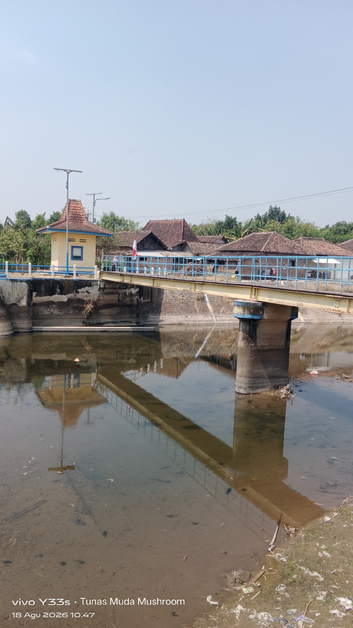 The image depicts a pedestrian bridge crossing a body of water in Tanjung, Indonesia. The bridge has a yellow walkway with a blue railing and is supported by concrete pillars. On the left side of the bridge, there is a small yellow building with a red-tiled roof, and behind it, a cluster of traditional Indonesian houses with red-tiled roofs. The water appears murky and shallow, with a sandy bottom visible in some areas. The reflection of the bridge and the surrounding buildings is visible on the water's surface. The scene is bathed in bright sunlight under a clear blue sky, suggesting a daytime setting. The image also includes a timestamp in the lower left corner: "18 Agu 2026 10.47".