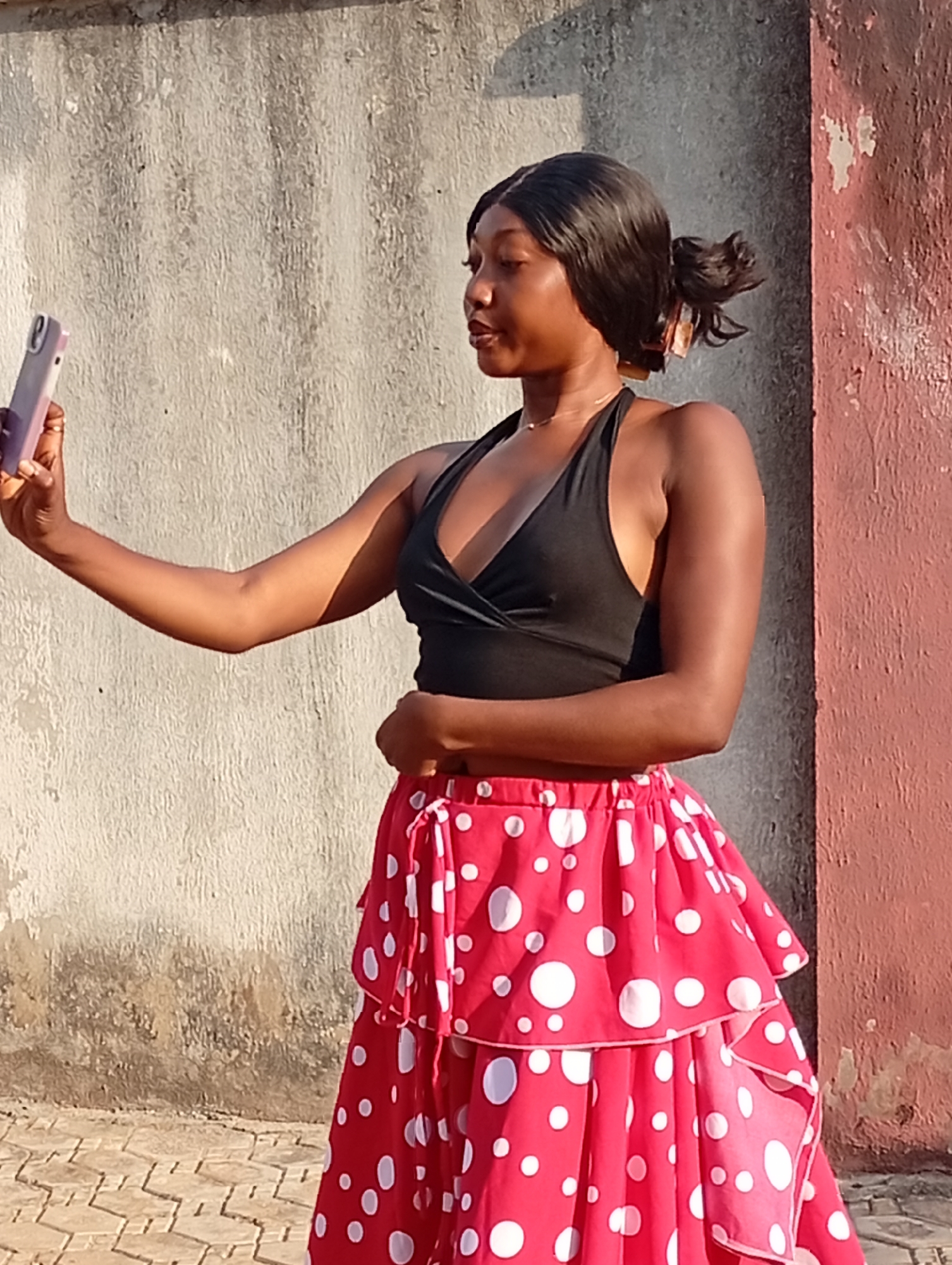 A young woman is taking a selfie outdoors. She is standing in front of a concrete wall that appears to be part of a building. The wall is a light grey color on the left side and transitions to a faded red on the right. The woman is wearing a black halter top and a red, layered skirt with white polka dots. Her hair is black and pulled back in a ponytail. She is holding a light-colored smartphone in her outstretched left hand. The ground is paved with patterned bricks. The location is Rido, Nigeria.