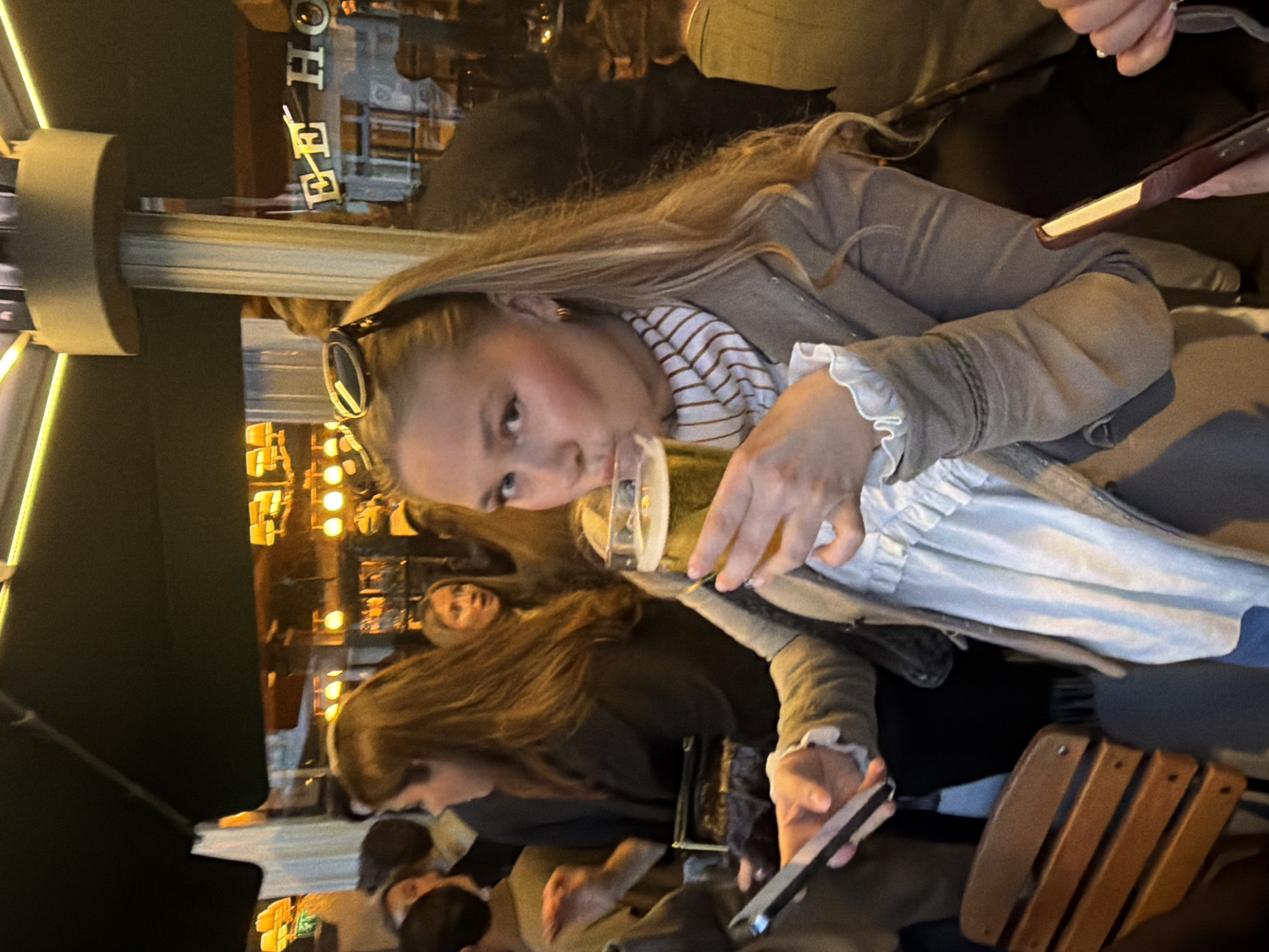The image captures a moment at an outdoor establishment, likely a bar or restaurant, in the city of London, United Kingdom. A young woman with long blonde hair is drinking from a glass of light-colored liquid, possibly beer. She is wearing sunglasses on her head, a striped shirt, a white blouse with ruffled cuffs, and a light brown jacket. Her posture suggests engagement with others at the table.
Other people are visible in the background and foreground, some holding smartphones. The environment includes elements of urban architecture, such as a green awning, streetlights, and signage that is in another language. The lighting suggests it is evening, as indicated by the dim ambient light and the illuminated building in the background. The foreground shows a wooden chair.