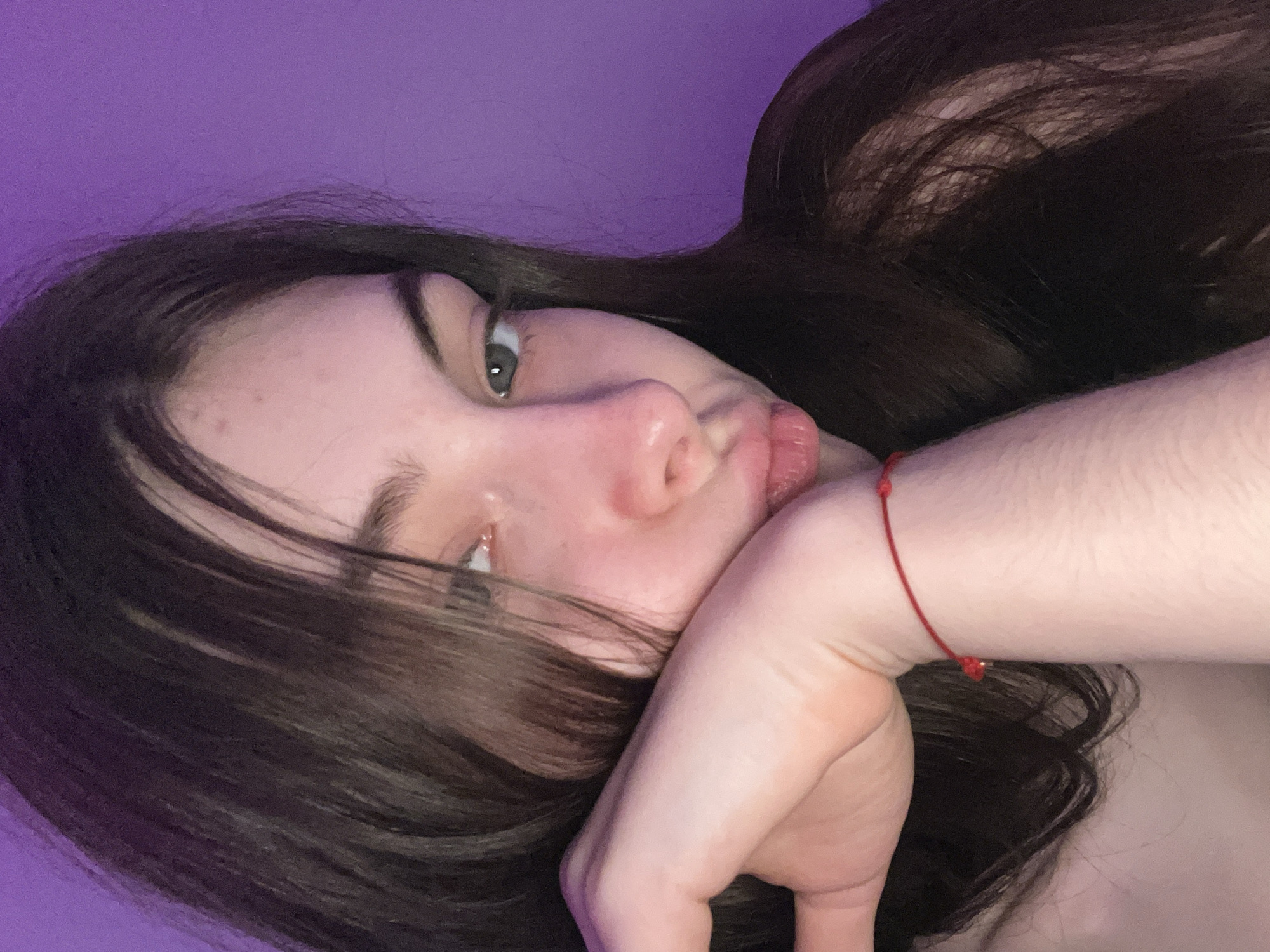 A young woman with long, dark hair is lying down, with her face resting on her hand. She is wearing a red bracelet on her wrist. The background is a solid purple color. The image appears to be taken from a close-up, low angle. There is no evidence of the city of Saint Petersburg, Russia.
