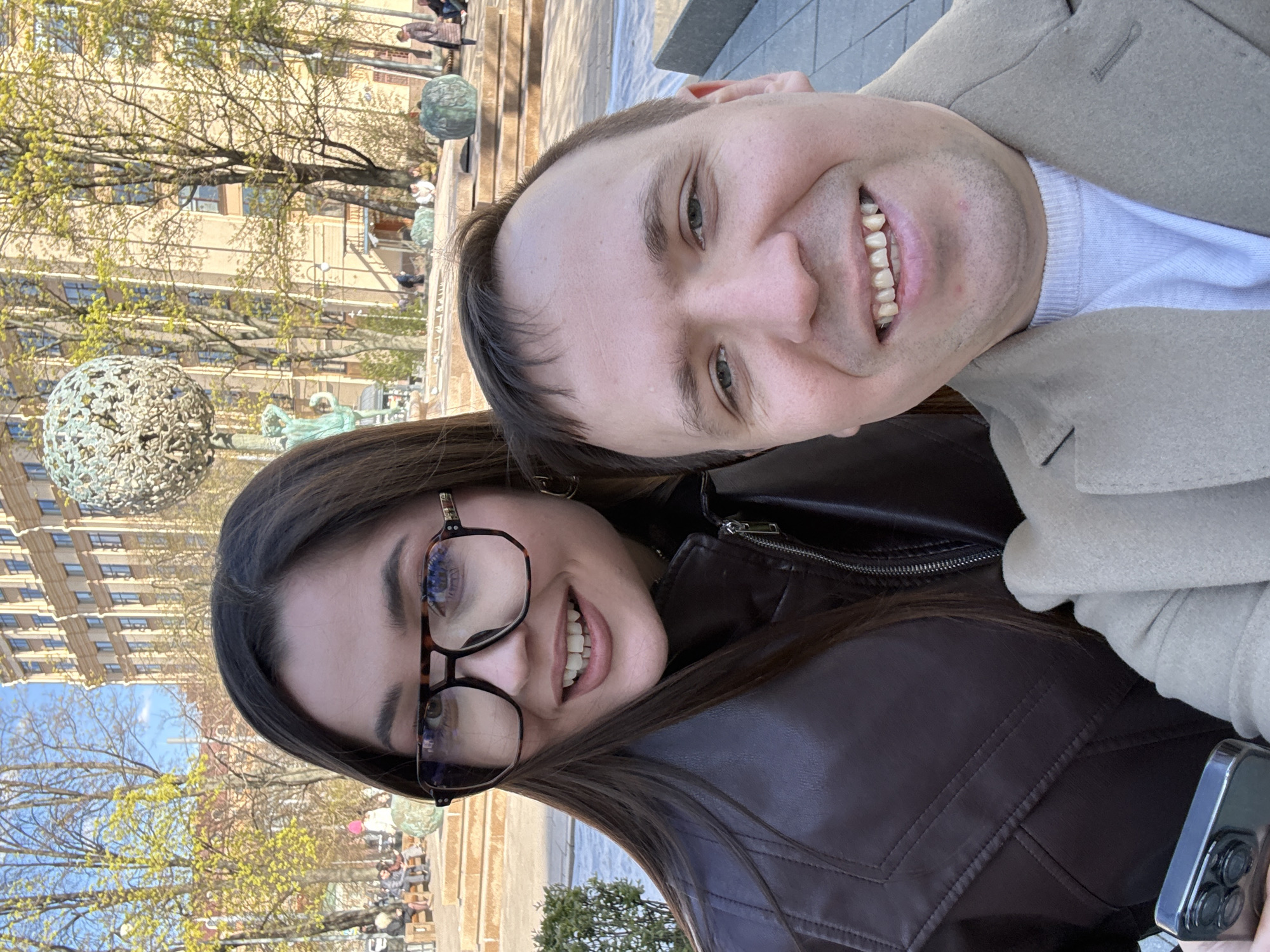 A close-up selfie of a man and a woman. The woman, on the left, has long dark hair, is wearing glasses, and a brown leather jacket. The man, on the right, is wearing a light tan coat over a white shirt. Both are smiling broadly, showing their teeth. The background shows an outdoor scene with a fountain, trees with budding leaves, and a building with many windows. Round decorative globes are visible on the fountain's edge. Other people are sitting on benches in the background. The photo appears to be taken during the day with natural lighting.