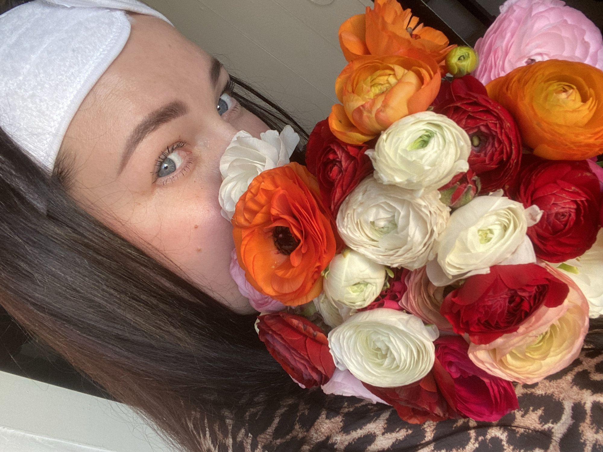 A close-up selfie shows a woman with fair skin, blue eyes, and dark brown hair. She is wearing a white headband. A bouquet of brightly colored ranunculus flowers, including orange, red, pink, and white varieties, is positioned near her face. Some of the flowers are close to her nose. The woman is wearing a brown and black patterned shirt or dress. The background is slightly out of focus and appears to be a light-colored wall or surface. The image does not provide enough information to determine a location in Sochi, Russia.
