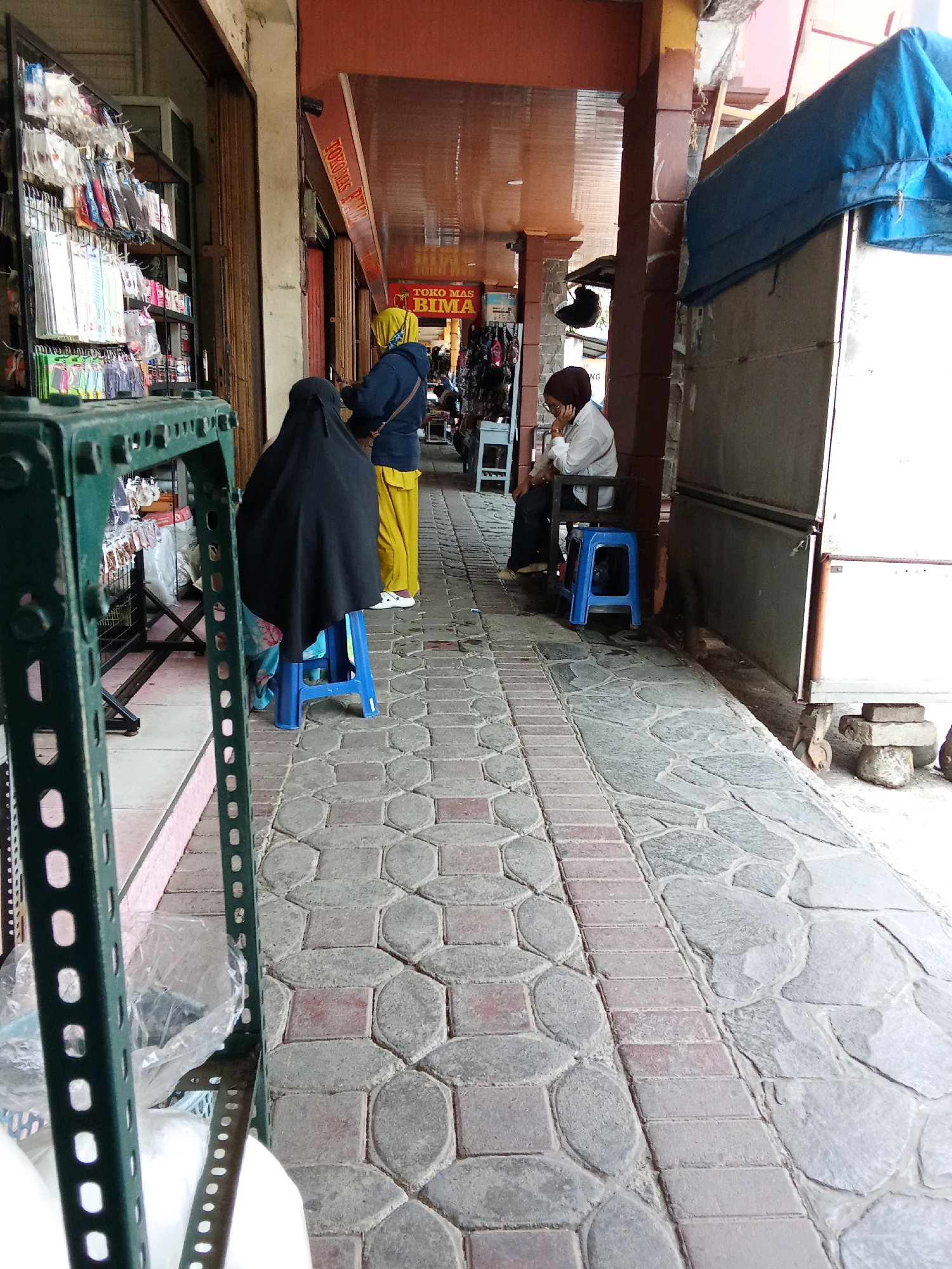 The image depicts a street scene in Banjarnegara, Indonesia, specifically along a pedestrian walkway in front of a row of shops. The walkway is paved with interlocking stones and accented with a strip of red bricks. In the foreground, a green metal shelving unit is visible. Further down the walkway, two individuals are seated. One, wearing a black hijab and covering, is on a blue plastic stool, with part of a blue and white patterned fabric visible beneath them. The second individual, wearing a white long-sleeved shirt, dark pants, and a brown hijab, is seated on a dark chair, also with a blue plastic stool beneath. Behind them, a shop sign reads "TOKO MAS BIMA". To the right of the walkway, a white kiosk or stall is partially covered with a blue tarp, and rests on concrete blocks and wheels. The environment appears to be an outdoor commercial area, likely a market or shopping street.