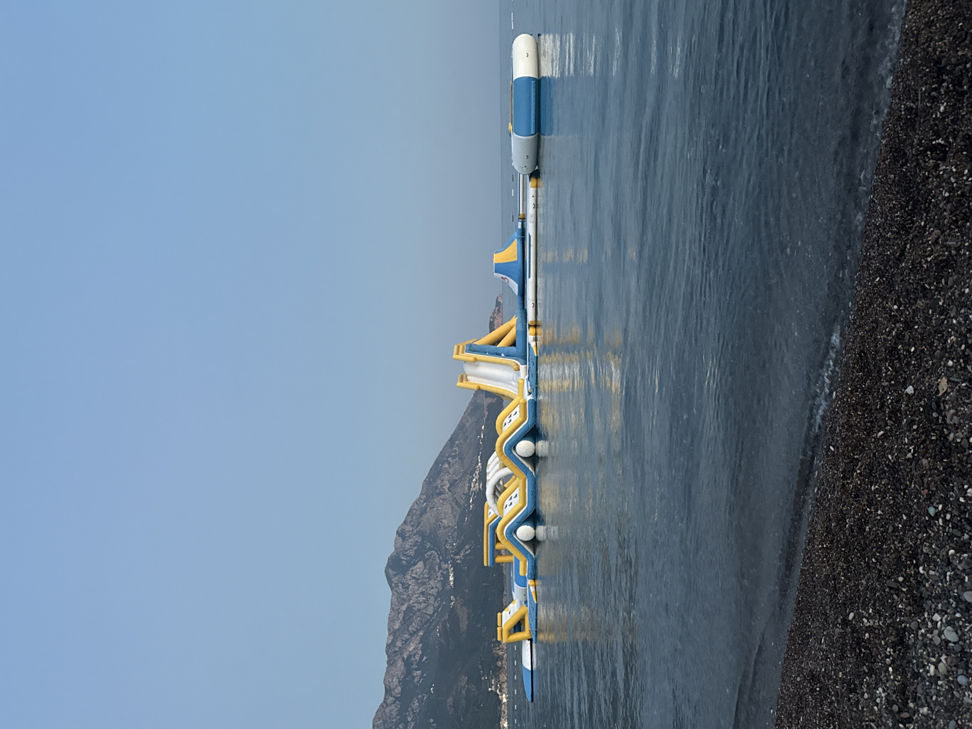 A long, inflatable water park with yellow and white slides and blue accents floats on the calm, dark blue water of the Adriatic Sea. The park is situated in front of a steep, green, and rocky mountainside dotted with white buildings. In the foreground, a dark, pebbly beach meets the water. The sky is a clear, light blue. The scene appears to be in Boreti, Montenegro.