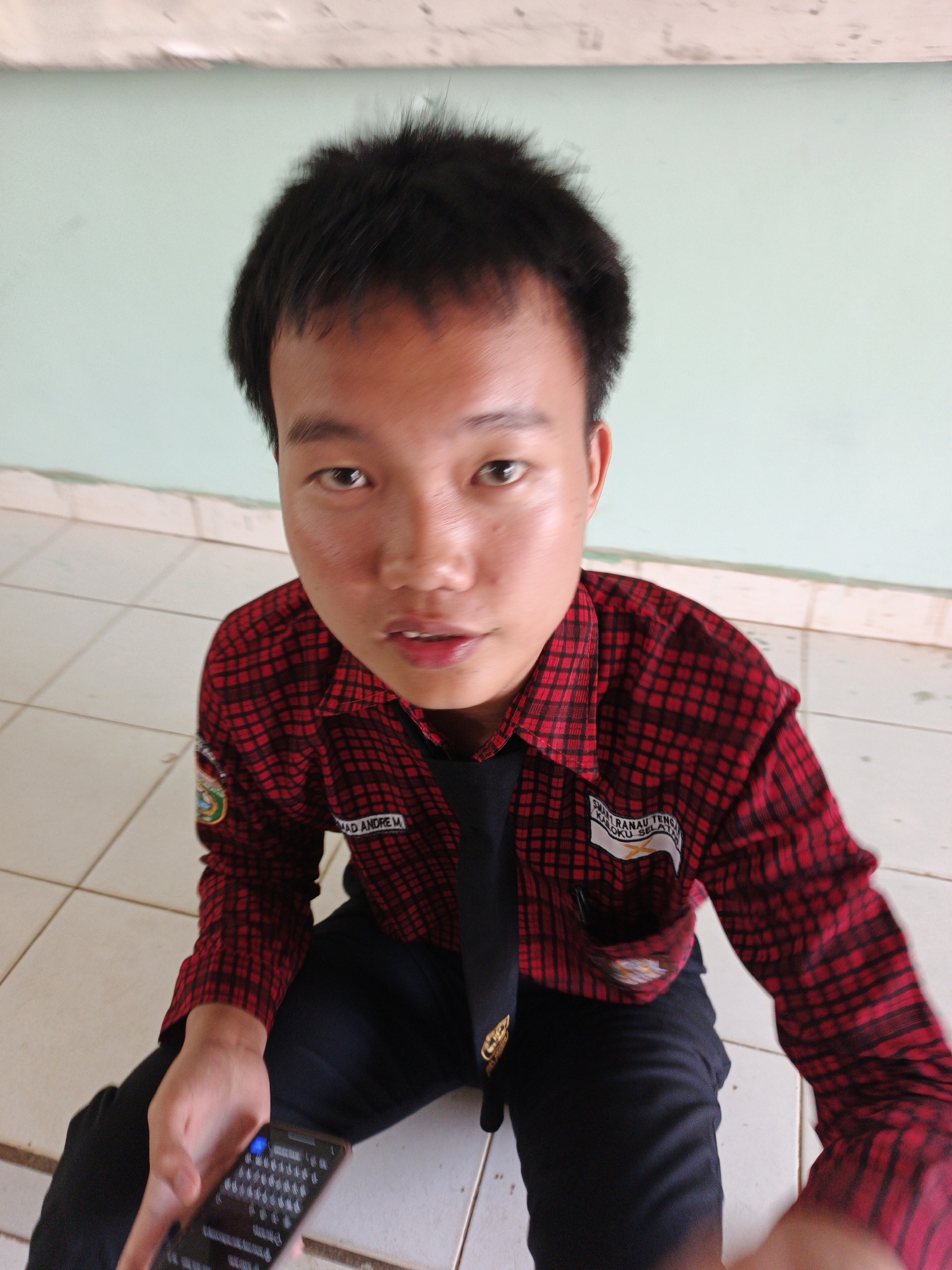 A young male with dark, spiky hair and brown eyes is seated on a tiled floor. He is wearing a red and black plaid collared shirt over a black t-shirt, with a black tie. The shirt has patches on the left breast and left sleeve. His right hand is holding a smartphone, the screen of which is displaying a keyboard. The background consists of a light green wall and a beige tiled floor. This scene is in the city of Tanjung Setia, Indonesia.