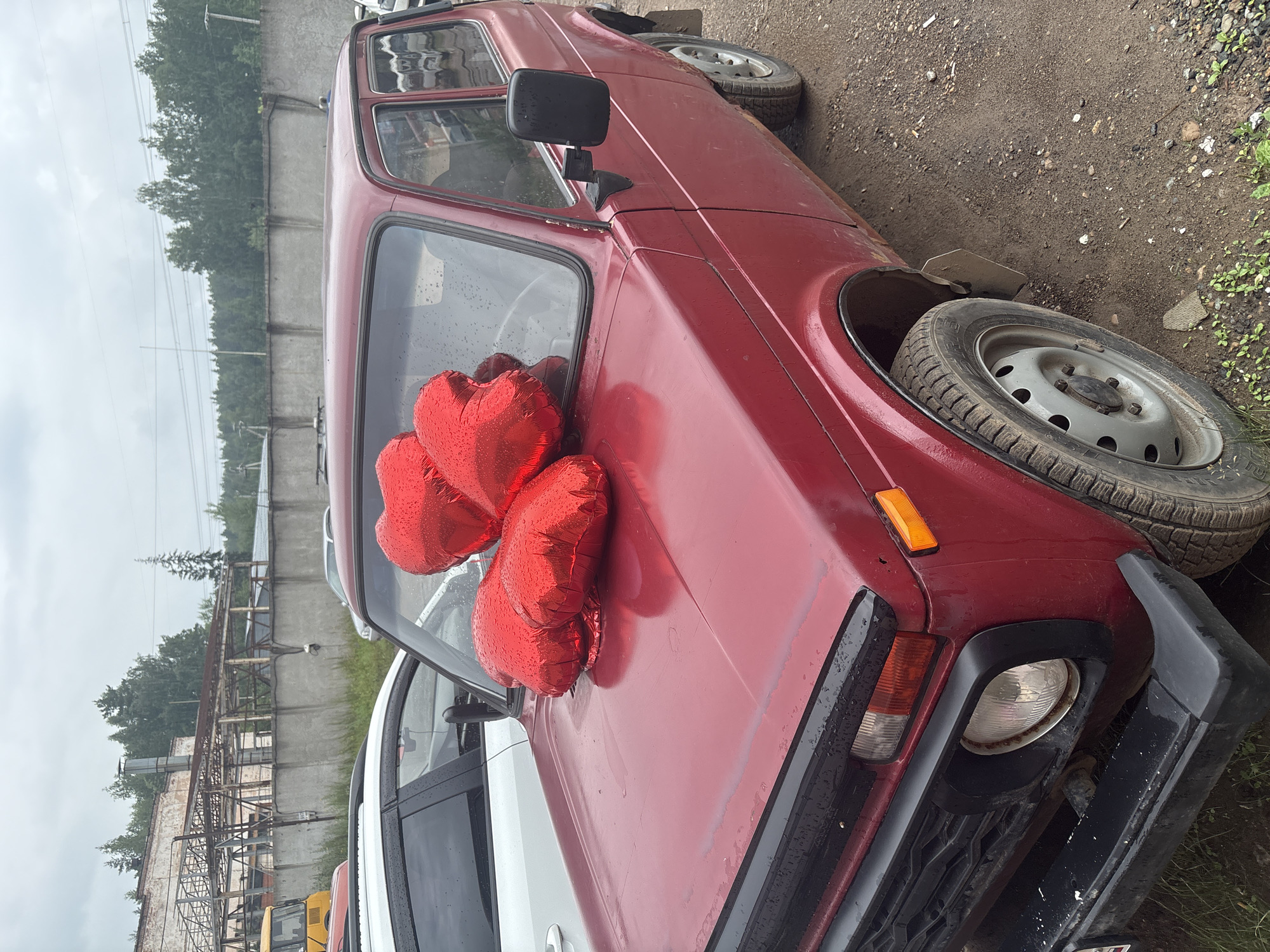 A red Lada Niva is parked in an outdoor lot in Noviy, Russia, on a cloudy day. Three red heart-shaped balloons are placed on the hood of the car, partially obscuring the windshield. The car appears to be older, with some visible rust and wear. A white car is parked to the left of the Lada, and a concrete wall with an industrial building structure behind it forms the background. The ground is a mixture of dirt and gravel, with some patchy green vegetation. The overall scene suggests a casual outdoor setting, possibly a parking area associated with a business or industrial site.