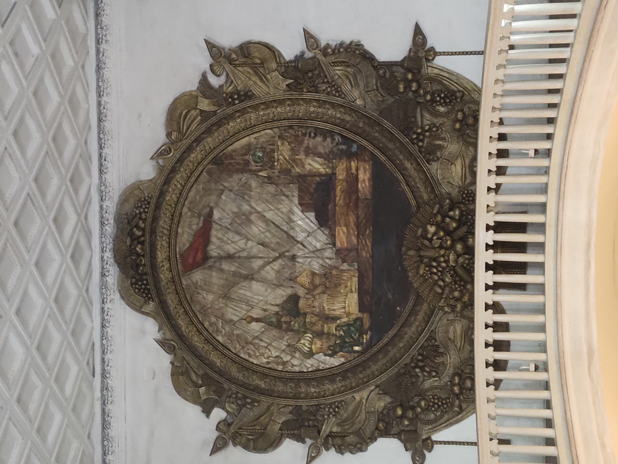 A circular painting hangs on a white wall in Moscow, Russia. The painting is encased in an ornate gold frame. The painting depicts a landscape with figures. A red flag is visible on the left side. The right side of the frame is partially obscured by a white slatted structure. The wall next to the painting has a diamond-patterned texture.