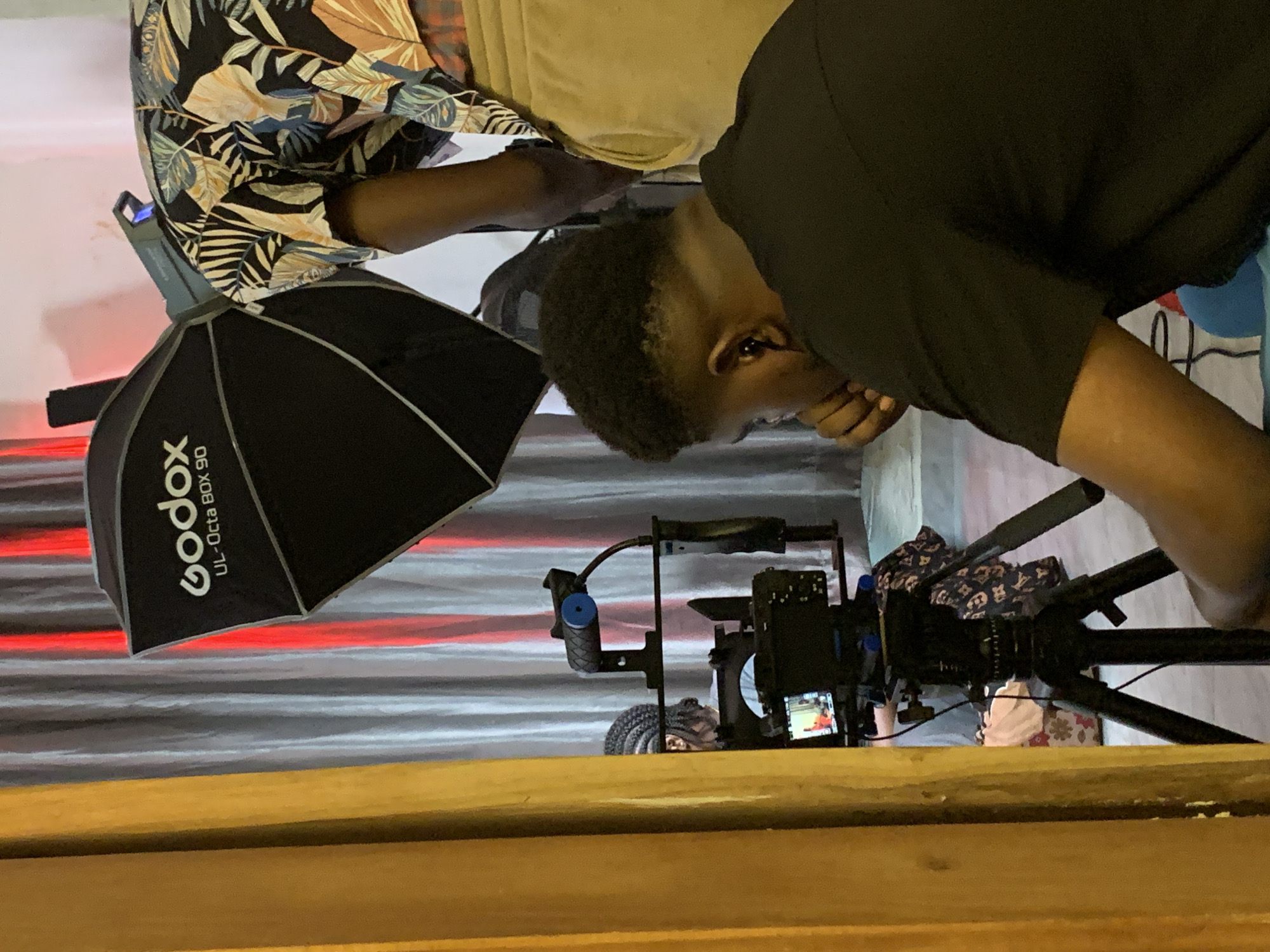An indoor, behind-the-scenes image captures a media production setup, likely in Ikorodu, Nigeria. The photograph shows two individuals working with professional camera and lighting equipment.
In the foreground, a man with short, dark hair, wearing a black t-shirt, is leaning forward, appearing to adjust or operate a camera rig. His expression is focused. Partially obscured behind him, another man wears a black shirt patterned with tropical leaves in blue, orange, and green.
Central to the scene is a professional camera rig, mounted on a tripod, featuring external handles. A small screen on the camera displays an active, brightly colored image, suggesting it is either recording or monitoring.
To the left, a "Godox UL-Octa Box 90" softbox is prominently displayed on a light stand. A light source atop this stand emits a blue glow, while another unseen light source casts a vivid red illumination across the background. The backdrop itself is a horizontally striped pattern in grey and white, interspersed with red stripes. A wooden surface runs across the bottom of the frame, possibly a counter or railing. Additional fabrics are visible near the tripod legs in the background.
The scene conveys a focused and productive environment dedicated to filming or photography, utilizing specialized equipment and artificial lighting.