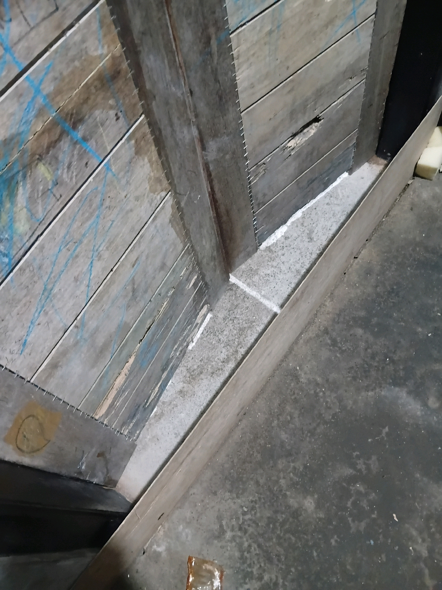 The image depicts a construction scene, likely in Cikal, Indonesia. In the foreground, a concrete floor with a grey, speckled texture is visible. A wooden plank, likely part of a temporary barrier or formwork, runs diagonally across the lower portion of the image. To the left, a section of a wooden wall constructed from horizontal planks is present. These planks are weathered and some show blue spray-painted markings. Small metal staples or nails are visible along the edges of the planks, suggesting assembly. A small section of what appears to be tape with a hand-drawn circular symbol is attached to one of the planks. In the background, a narrow channel filled with a light-colored material, possibly concrete or grout, separates the wooden wall from another dark vertical surface. A small, crumpled plastic bag is on the concrete floor in the lower right corner. The overall scene suggests ongoing construction or repair work.
