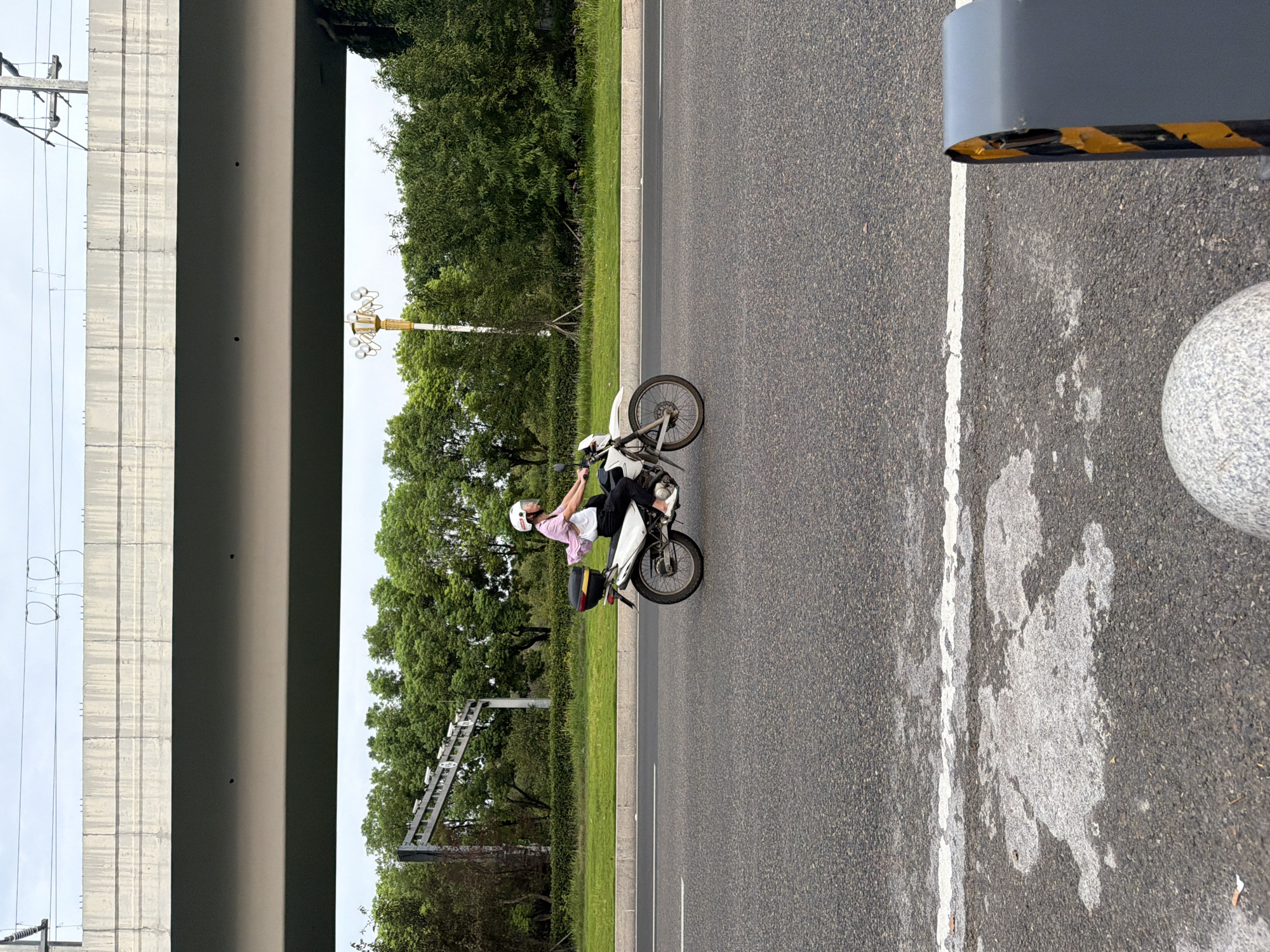 In Jiangjin, China, a person wearing a pink shirt, black pants, and a white helmet rides a white motorcycle on an asphalt road. The motorcycle has a black storage box on the back. In the background, there are green trees and a large concrete overpass with overhead wires. A yellow lamp post with multiple bulbs stands to the right of the motorcycle. The road has white lane markings and some discolored patches. In the foreground, part of a gray spherical object and a black and yellow striped bollard are visible. The sky is overcast.