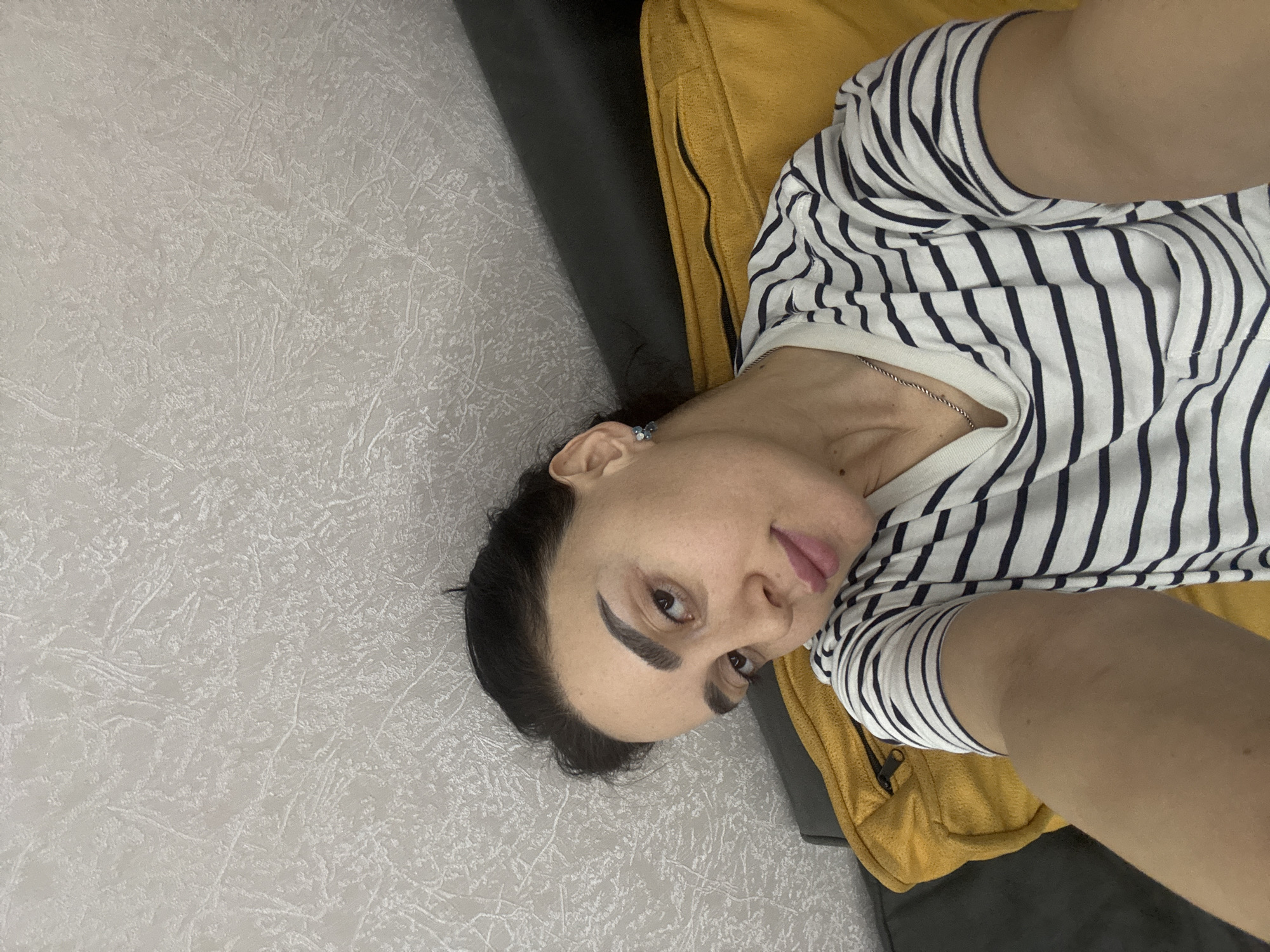 A woman is lying down, likely on a sofa or bed, wearing a striped shirt. She is looking at the camera. The background includes a textured wall, a yellow cushion, and a dark, possibly leather, part of the furniture. It is not possible to determine the location as Tbilisi, Georgia from the image content.