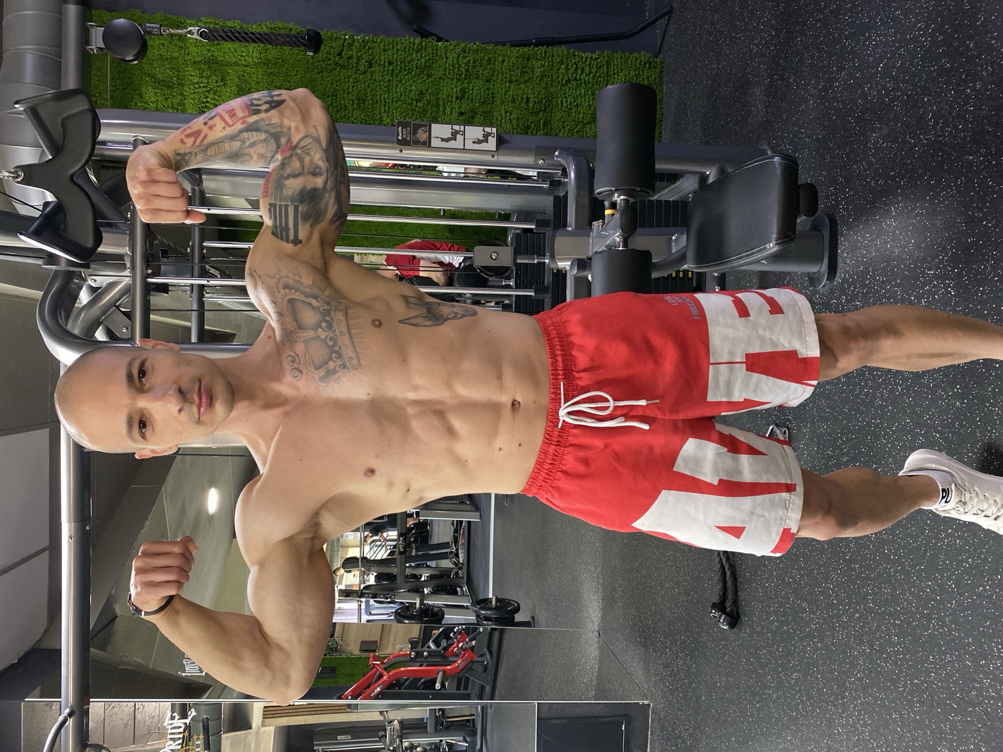 A muscular man is posing at a fitness center in Pleven, Bulgaria. He has a shaved head and fair skin, with numerous tattoos on his left arm and chest. He is wearing red and white shorts and white sneakers. He is flexing his arms, emphasizing his biceps and abdominal muscles. The background shows various gym equipment, including a cable machine, weights, and other exercise machines. The floor is dark with light speckles.