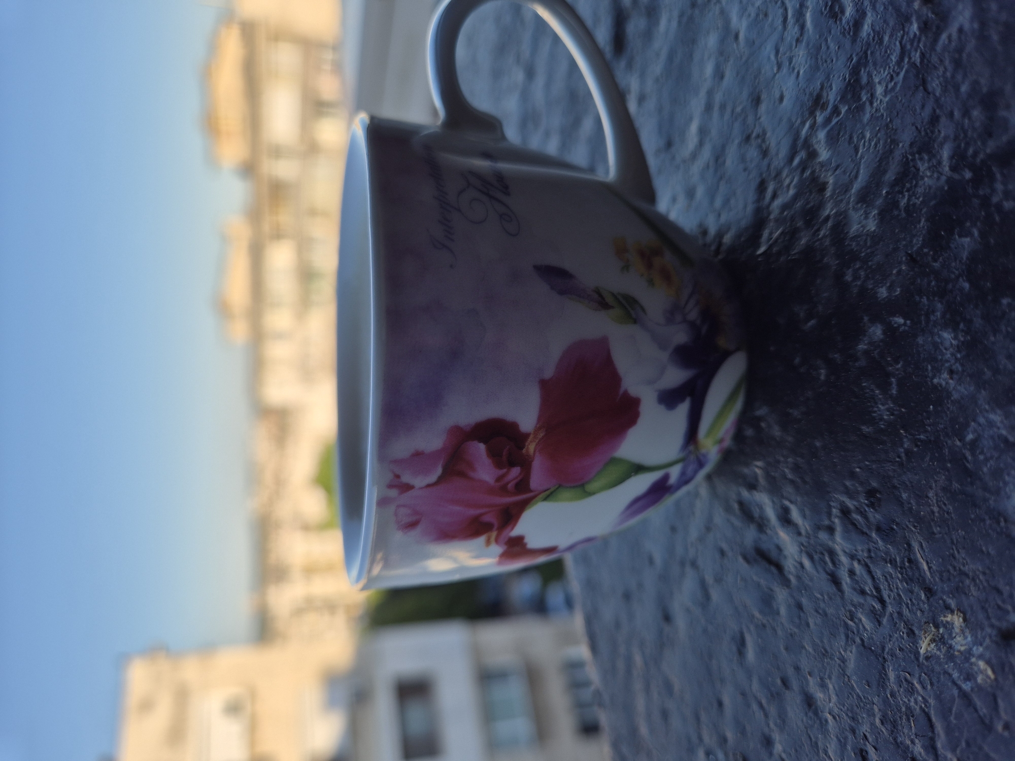 A close-up, low-angle outdoor shot shows a white coffee cup with a floral design, featuring a large pink iris and smaller yellow flowers and green leaves. The cup sits on a dark, textured surface, possibly a balcony railing, with a blurred cityscape in the background under a clear blue sky. Buildings with windows and balconies are visible, along with hints of trees and possibly a street. The lighting suggests it is either early morning or late afternoon.