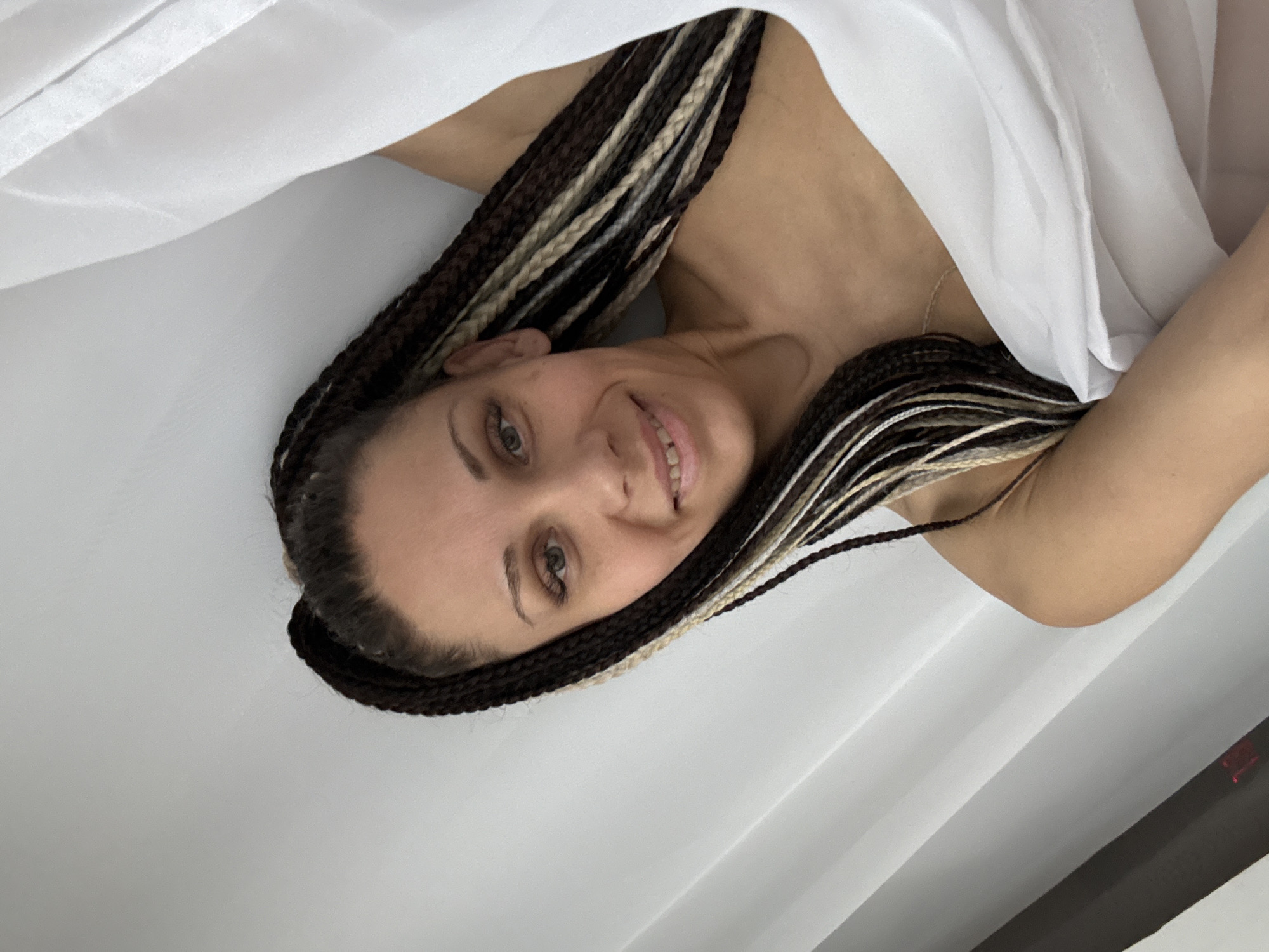 A woman with braided hair, featuring brown and blonde strands, smiles towards the camera. She is partially covered by a white cloth. The background is a white wall with light reflecting across it. The location cannot be determined.