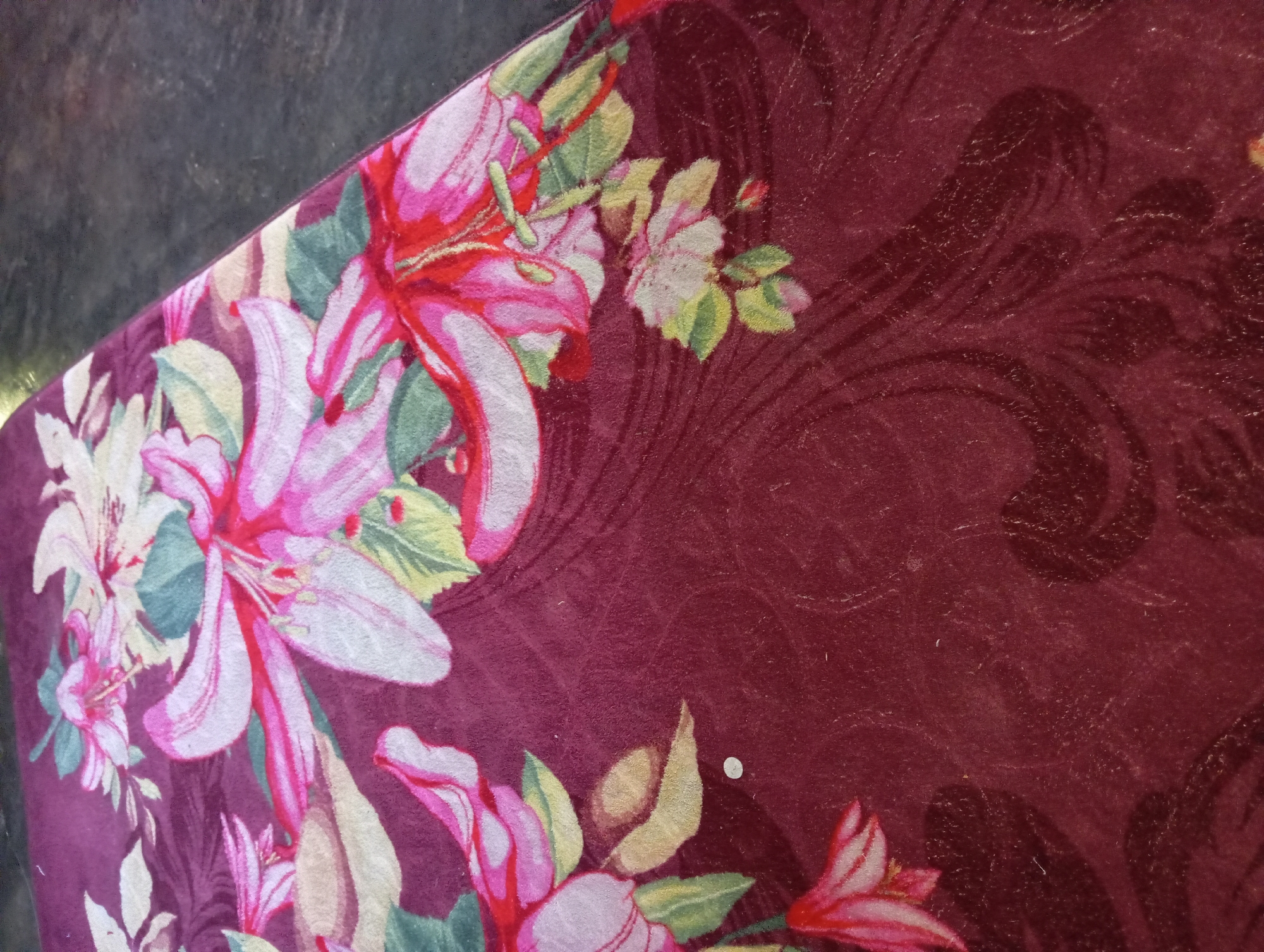 The image shows a close-up of a patterned rug. The rug has a deep maroon background with a floral design featuring large, stylized flowers in shades of pink, white, and yellow, with green leaves. The flowers appear to be lilies or similar trumpet-shaped blooms. Swirling, darker patterns are visible in the background of the rug. The rug is partially laid out on a dark, textured surface, which appears to be a floor. One corner of the rug is visible, suggesting it is placed in a room. The location in Rejosari, Indonesia, is inferred from context not present in the image itself, but the rug's pattern and material suggest it could be in a residential or commercial setting.