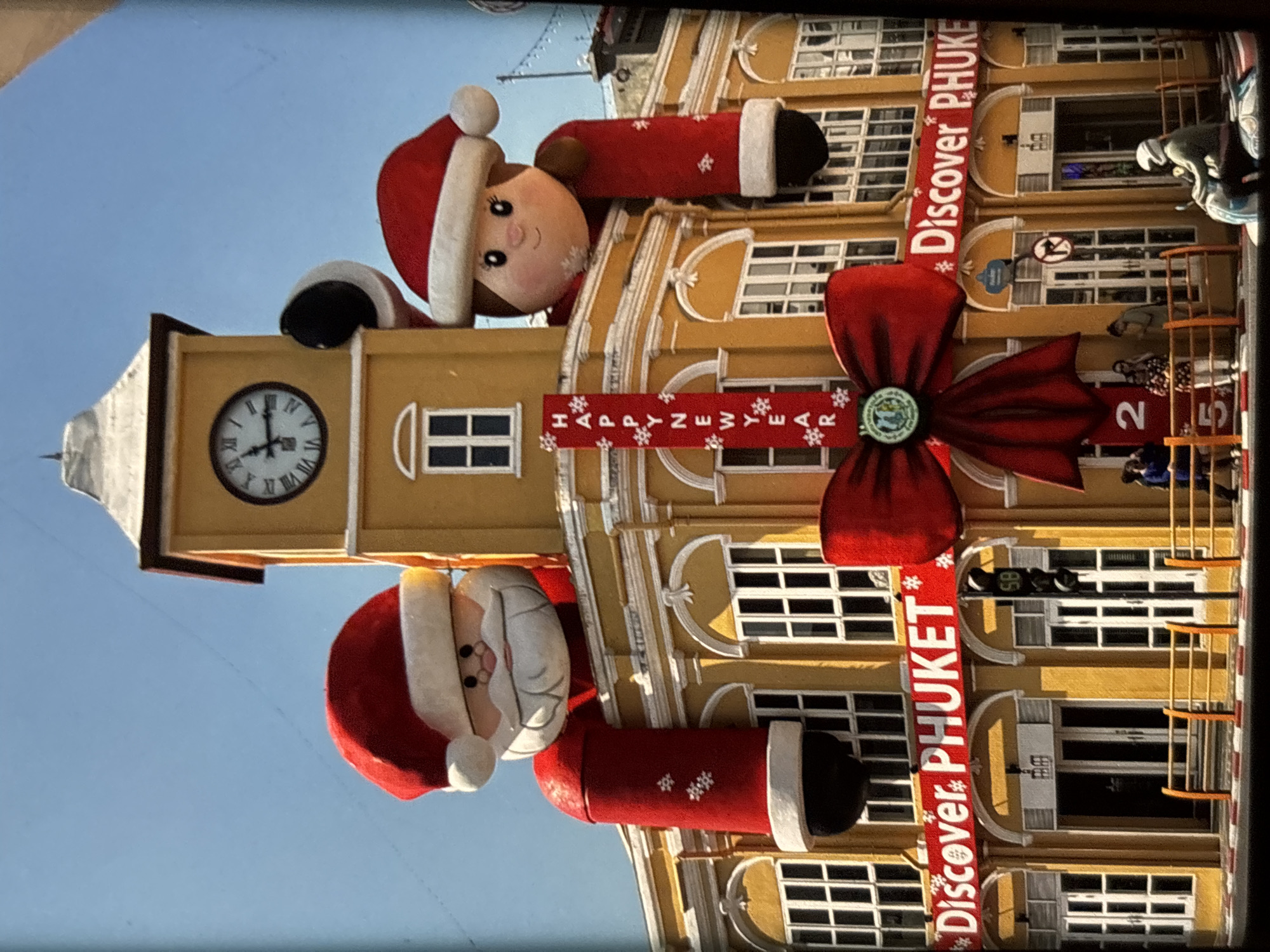 The image shows a festive building facade in Chalong, Thailand, decorated for the holiday season. The building is yellow with white trim and features large inflatable figures of Santa Claus and a child in Santa hats on either side of a clock tower. The clock tower is yellow with a white roof and a black clock face. A large red bow is draped across the lower part of the building, partially obscuring a banner that reads "Discover PHUKET". A vertical banner with "HAPPY NEW YEAR" is also visible. Several people are standing on the sidewalk in front of the building, some looking at the decorations. A motorcycle is parked on the right side of the frame, with a rider wearing a helmet. The sky is clear and blue. The overall scene suggests a public celebration or event.