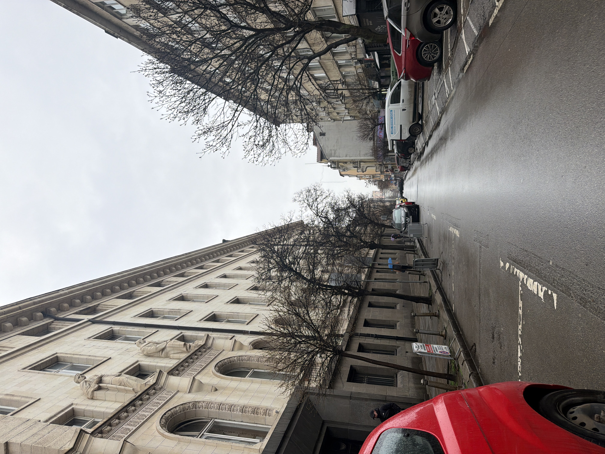 The image depicts a street scene in Sofia, Bulgaria, taken on a wet, overcast day. The road surface is wet and reflects light. Lined along the street on both sides are parked cars, including red, white, and dark-colored vehicles. Buildings flank both sides of the road, with the structure on the left exhibiting classical architectural features, including sculpted figures on its facade. Bare trees with leafless branches line the street between the buildings. Pedestrians are present on the sidewalk, including one person near the bottom right corner of the image. A street sign is visible attached to the building on the left. The overall impression is of a city street on a typical day.