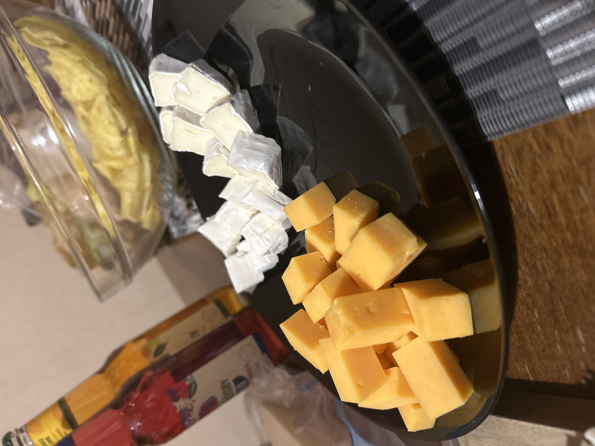 This close-up image captures a platter of cubed cheeses, likely prepared for a snack or appetizer. On a dark, highly reflective surface, two distinct types of cheese are arranged. To the left, numerous cubes of a light-colored, off-white cheese with a visible soft rind, possibly Brie or Camembert, are piled. To the right, a generous heap of bright yellow or orange cheese cubes, resembling cheddar or a similar hard cheese, is present. The reflective surface of the platter shows blurred reflections of the surroundings.
Partially visible on the far left, a clear glass bowl contains a quantity of light yellow, shredded or stringy food, whose exact nature is unclear. Below the platter, two bottles are visible, with their labels partially showing colorful designs in yellow/orange and red/pink, but no discernible text. The background features a patterned mat or placemat in grey tones behind the platter, and a wooden surface, likely a table or countertop, along the right side of the frame. The scene is indoors, illuminated by artificial light, suggesting an evening or late afternoon setting. There are no people visible, and the overall impression is one of casual food preparation or serving in a home environment in Sofia, Bulgaria.
