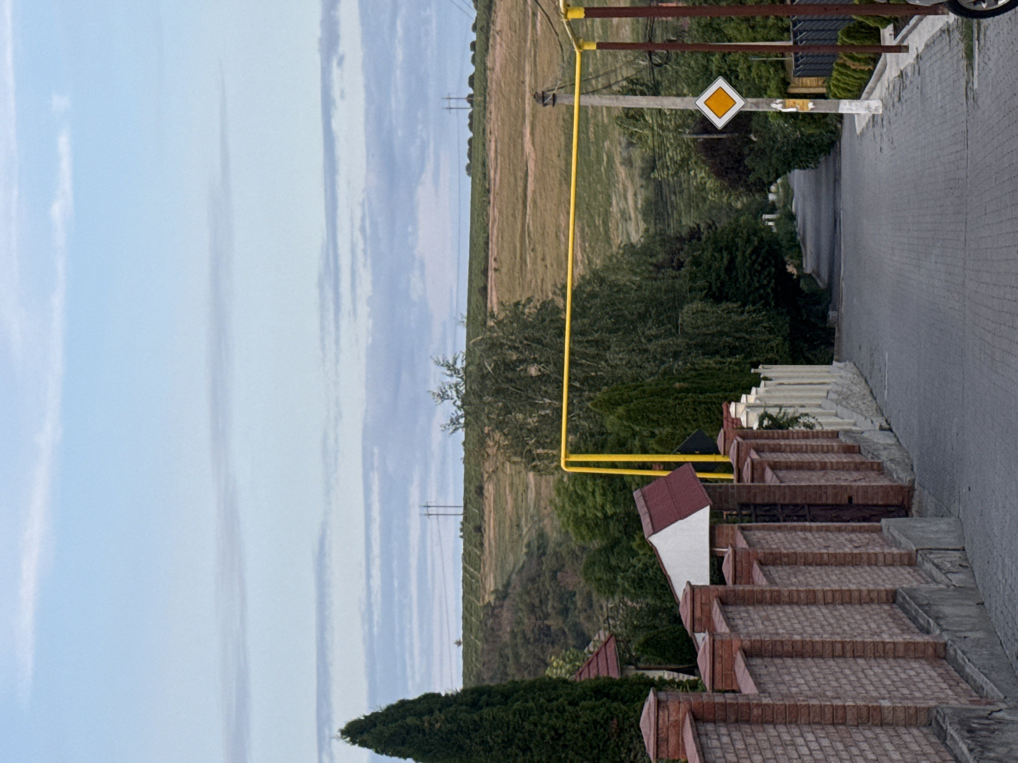 The image depicts a straight, paved road in Ialoveni, Moldova, lined with a brick fence on the left and various trees and vegetation on the right. In the distance, a gently sloping hill is visible, covered with fields and dotted with power lines. The sky is partly cloudy. A yellow gas pipe runs horizontally across the middle ground of the image, supported by poles. A diamond-shaped road sign with an orange border and white interior is mounted on a utility pole on the right side of the road. The overall scene suggests a suburban or semi-rural residential area.