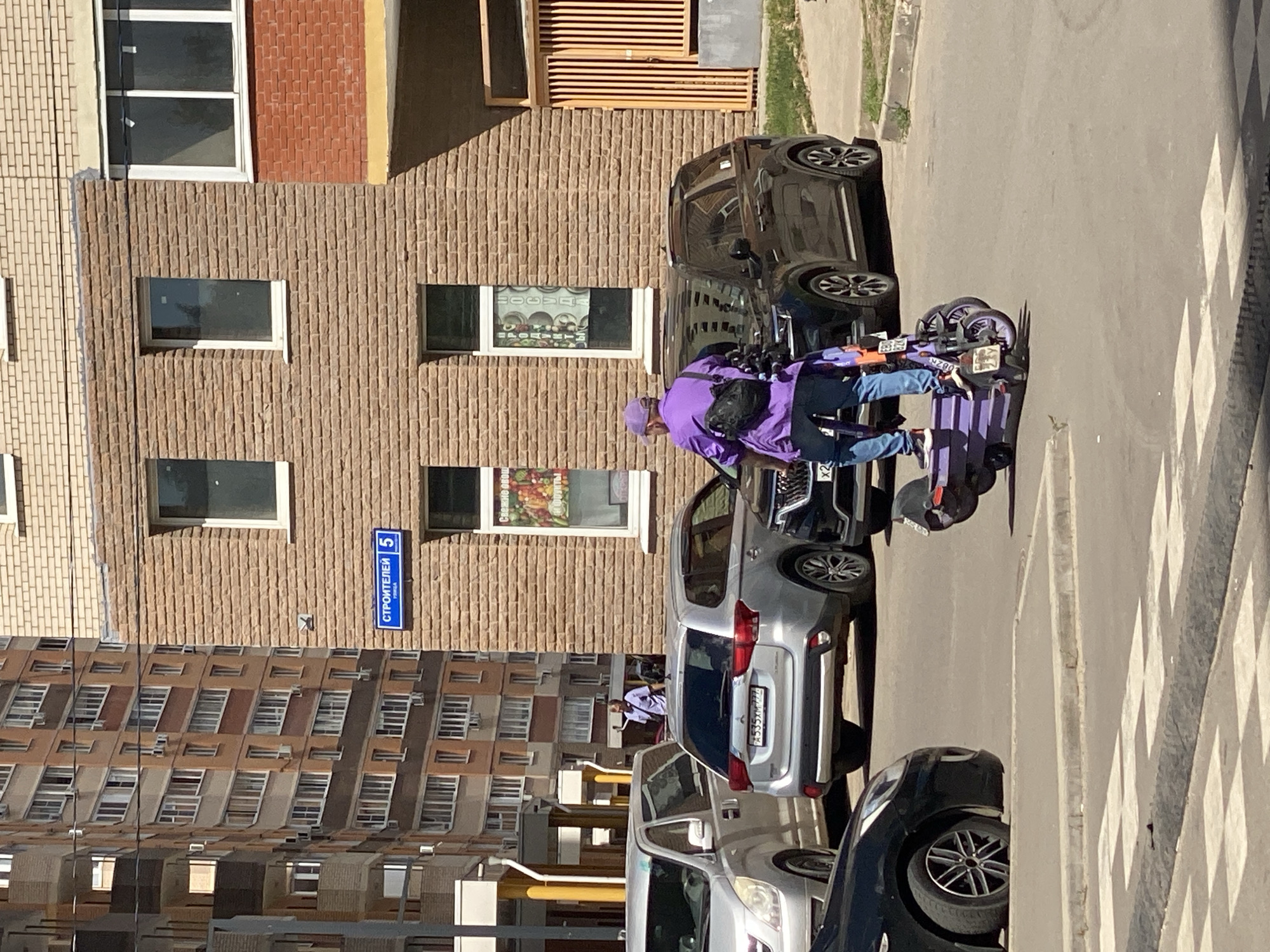 In Kotelniki, Russia, under the bright daylight and clear weather, a person in a vibrant purple jacket and matching hat is on an electric scooter. The scooter itself is also a striking purple, parked on a paved street marked with a pedestrian crossing. The scene is set against a backdrop of a multi-story building with a facade of light brown brick, featuring numerous windows, some with white frames, and others with darker glazing. To the left, another building with a similar brick pattern is visible. Several cars are parked along the street; a dark-colored car is in the foreground, and a silver SUV is parked behind it. A blue street sign with white text, reading "СТРОИТЕЛЕЙ 5," indicating the street name and building number, is affixed to the brick building. The overall impression is of an everyday urban scene in a residential area.