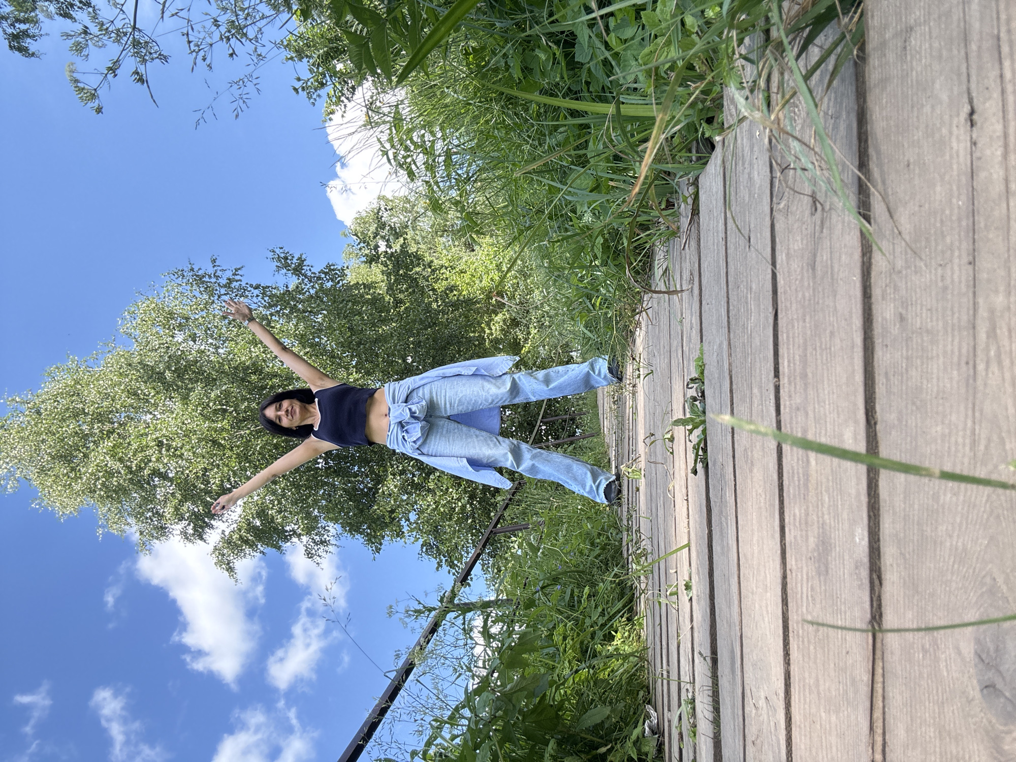 An adult woman with dark hair and fair skin is standing on a wooden bridge with her arms raised in the air. She is wearing a dark blue tank top, light blue jeans, and a light blue unbuttoned shirt tied around her waist. The bridge is surrounded by lush green vegetation and tall trees. The sky is blue with scattered white clouds. The scene appears to be an outdoor natural setting. The location is the city of Chaikovsky, Russia.