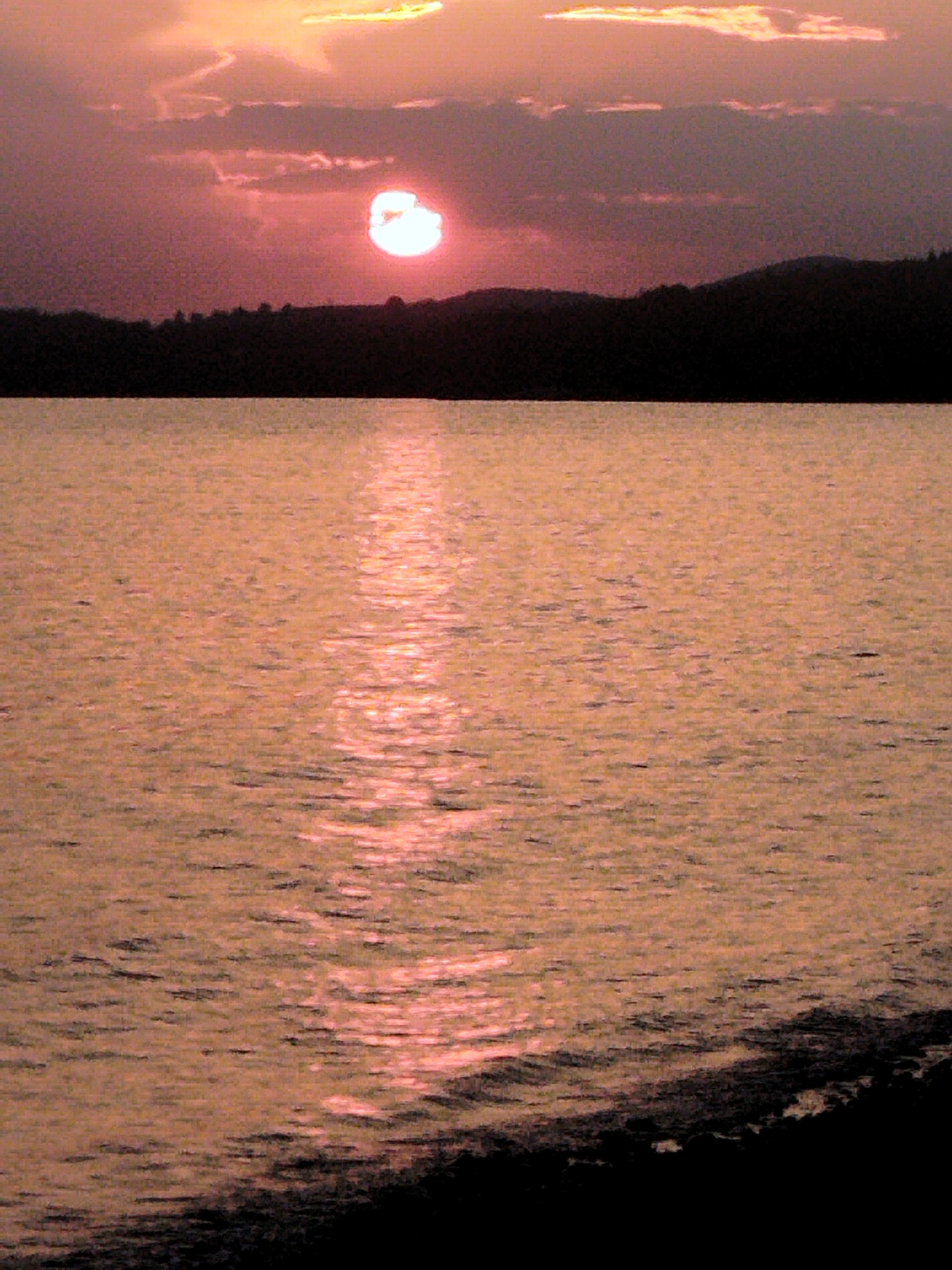 A sunset over a body of water is depicted. The sun, a bright, hazy orb, is low in the sky, casting a warm, pinkish-orange glow across the scene. Clouds, tinged with the same colors, are scattered above. The water is calm, reflecting the sunset with a shimmering path of light leading towards the viewer. The horizon is marked by a dark, silhouetted line of trees or hills, suggesting a natural landscape. The bottom edge of the image shows a dark, indistinct foreground, possibly land or the edge of the water body. The location is the Sithonia Municipal Unit in Greece, a coastal region known for its natural beauty and sunsets over the Aegean Sea.