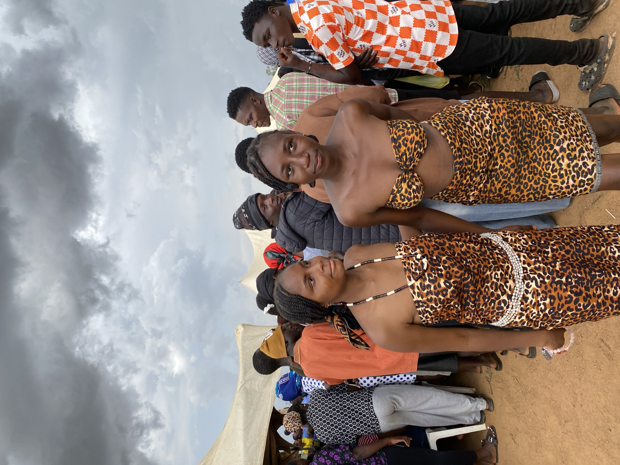 A group of people stand outdoors under a cloudy sky in what appears to be Durimi, Nigeria. Two women in the foreground wear leopard print dresses. Behind them are other individuals in various attire, including casual shirts and dresses. The setting includes a tent structure on the left and what appears to be a dirt ground. The people seem to be gathered, though their specific activity is unclear.