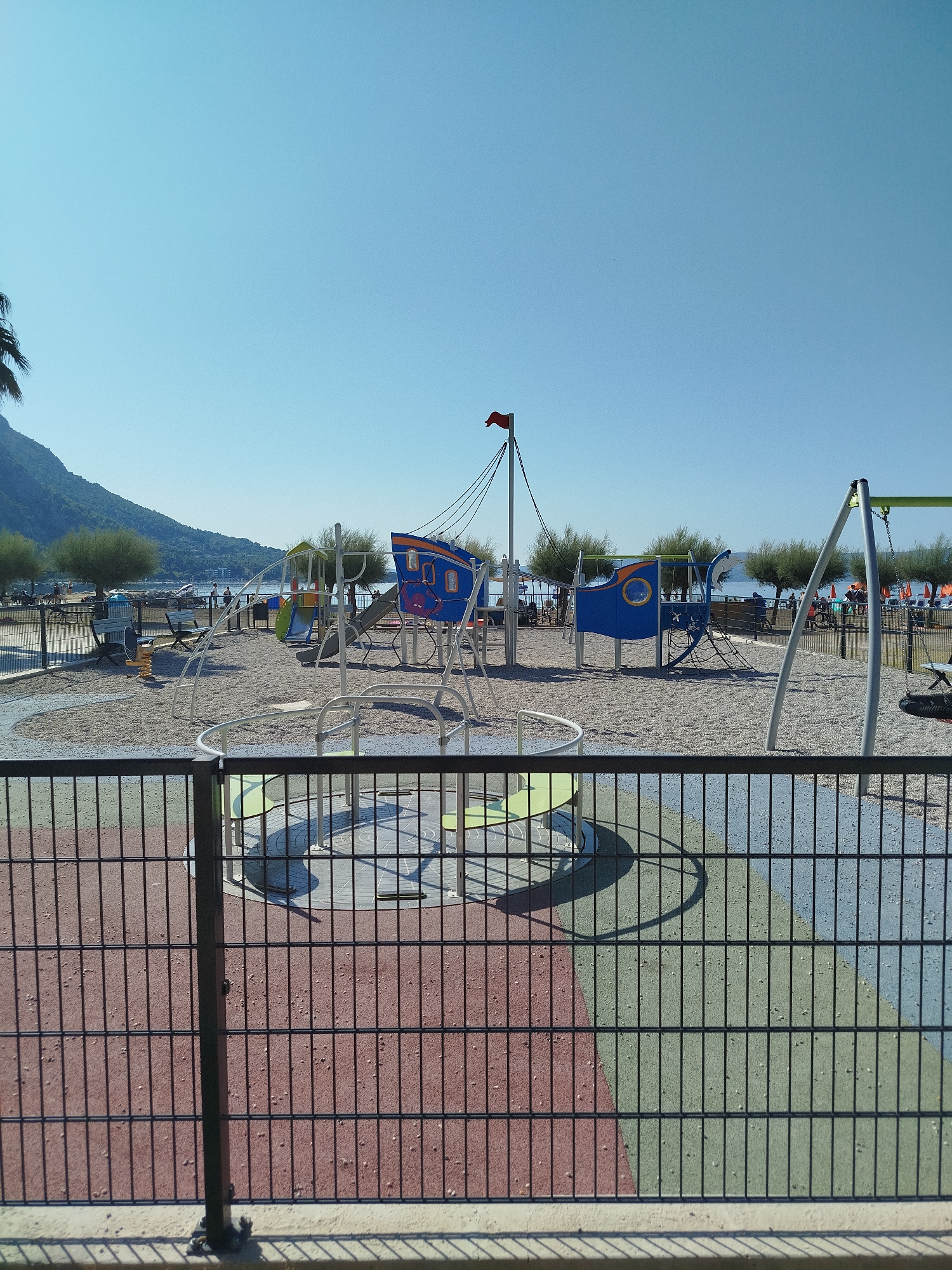 A sunlit, empty playground is depicted near the coast in Omiš, Croatia. The playground features a large, blue, boat-shaped play structure with orange accents, a slide, and a rope climbing net. To the left of the boat structure is a smaller blue play element. Further to the left, a yellow spring rider is visible. White metal frames for swings are present on the right side of the playground, and a merry-go-round with colorful rubberized surfacing in red, green, and blue is partially visible in the foreground behind a dark metal fence. The ground of the playground is covered in light-colored gravel.
In the background, a body of water, likely the sea, is visible, with distant hills or mountains on the left side of the frame. A palm tree is partially visible on the far left, and a red flag flies from a mast on the boat structure. A few people are visible in the distance near the water and beach, some appearing to be on bicycles, and several orange beach umbrellas are set up. The sky is clear and blue. A dark metal fence runs across the bottom of the image, separating the viewer from the playground.