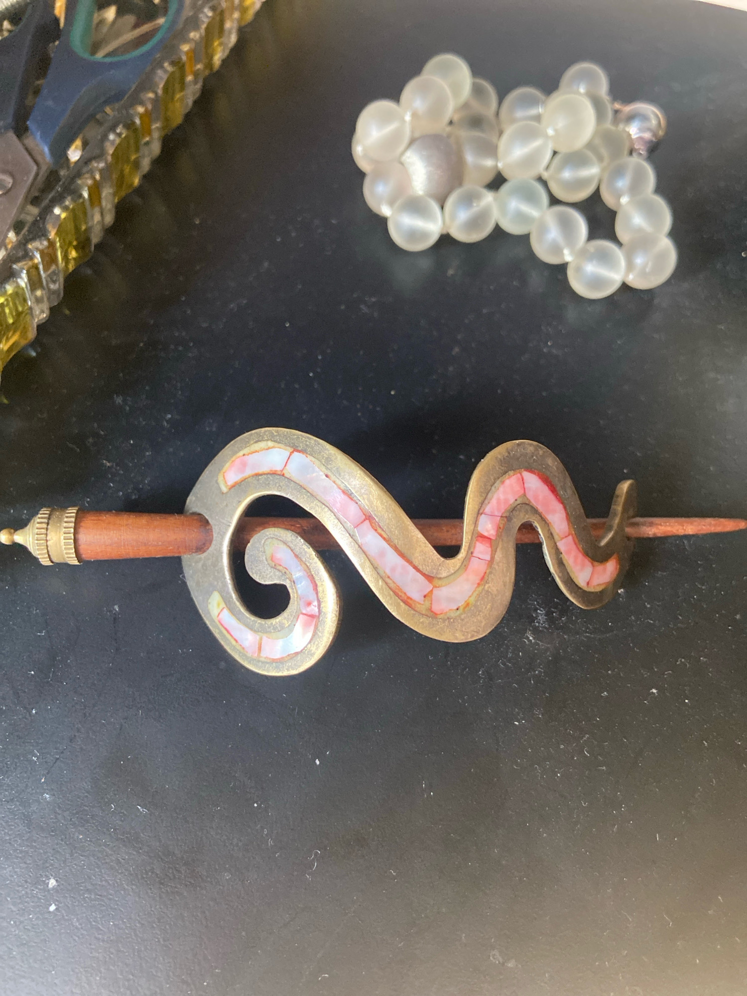 The image shows a close-up view of a decorative hair stick with a coiled, metallic design featuring inlays of pink and white material, possibly shell or mother-of-pearl. The hair stick is threaded through a wooden dowel. In the background, to the left, is the edge of a pair of scissors with yellow accents, and to the right, a strand of translucent, bead-like objects. The surface appears to be a dark, possibly textured material. There is no information in the image to suggest a location in Kassel, Germany.