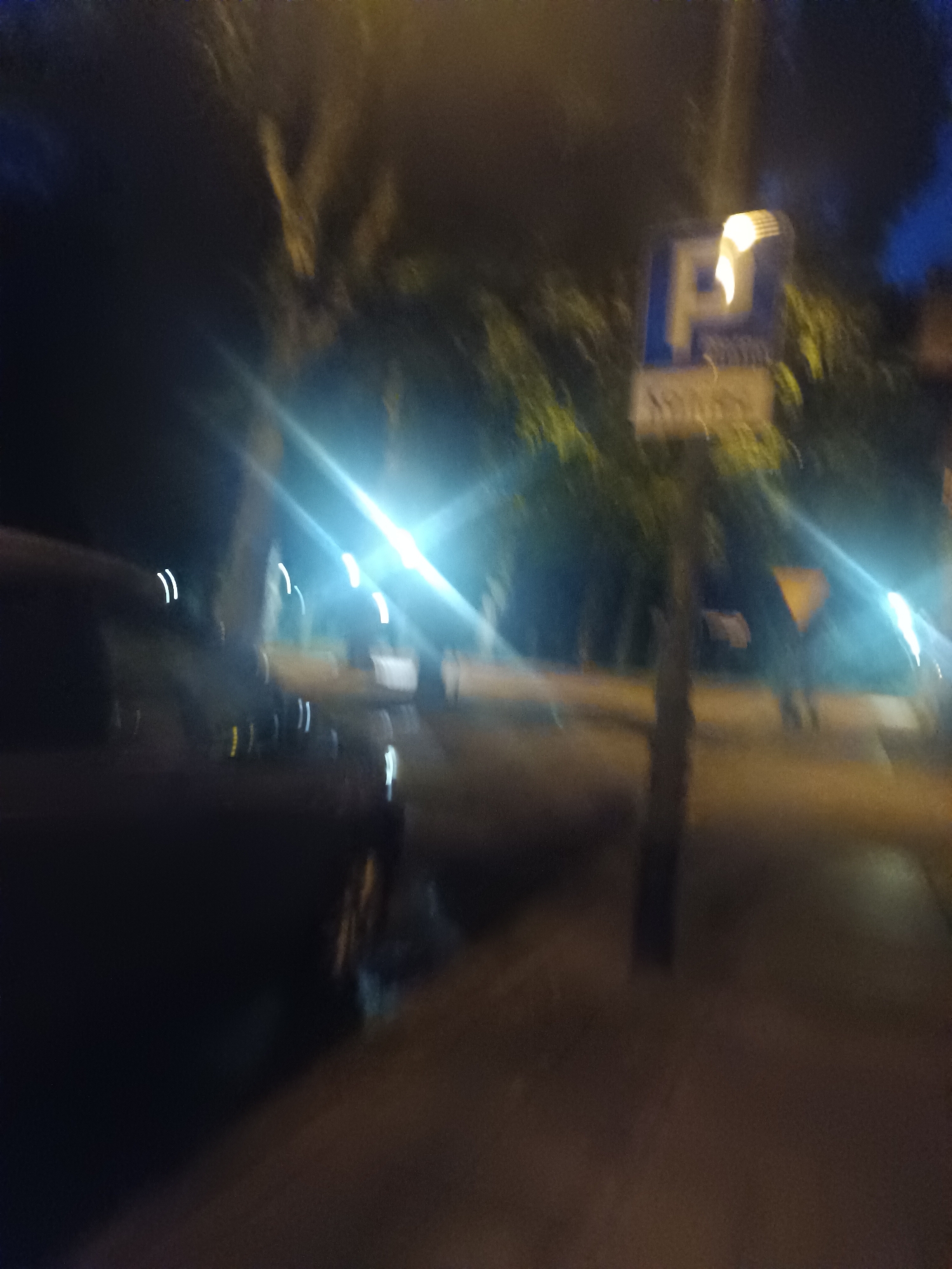 The image captures a nighttime scene on a street in Świnoujście, Poland, likely during the late evening or early morning. A dark-colored vehicle is parked on the left side of the frame, its shape blurred by low light conditions and motion. In the background, a street sign is visible, displaying a blue square with a white "P" symbol, indicating parking. Above the "P," there appears to be a yellow, curved element, possibly a light or reflection. Below the parking symbol, there is another sign with text, which is illegible due to blurriness. The street is illuminated by artificial lights, creating bright streaks and lens flares that distort the view of the surroundings, including trees and possibly other street elements. The overall impression is that of a quiet, dimly lit urban environment.