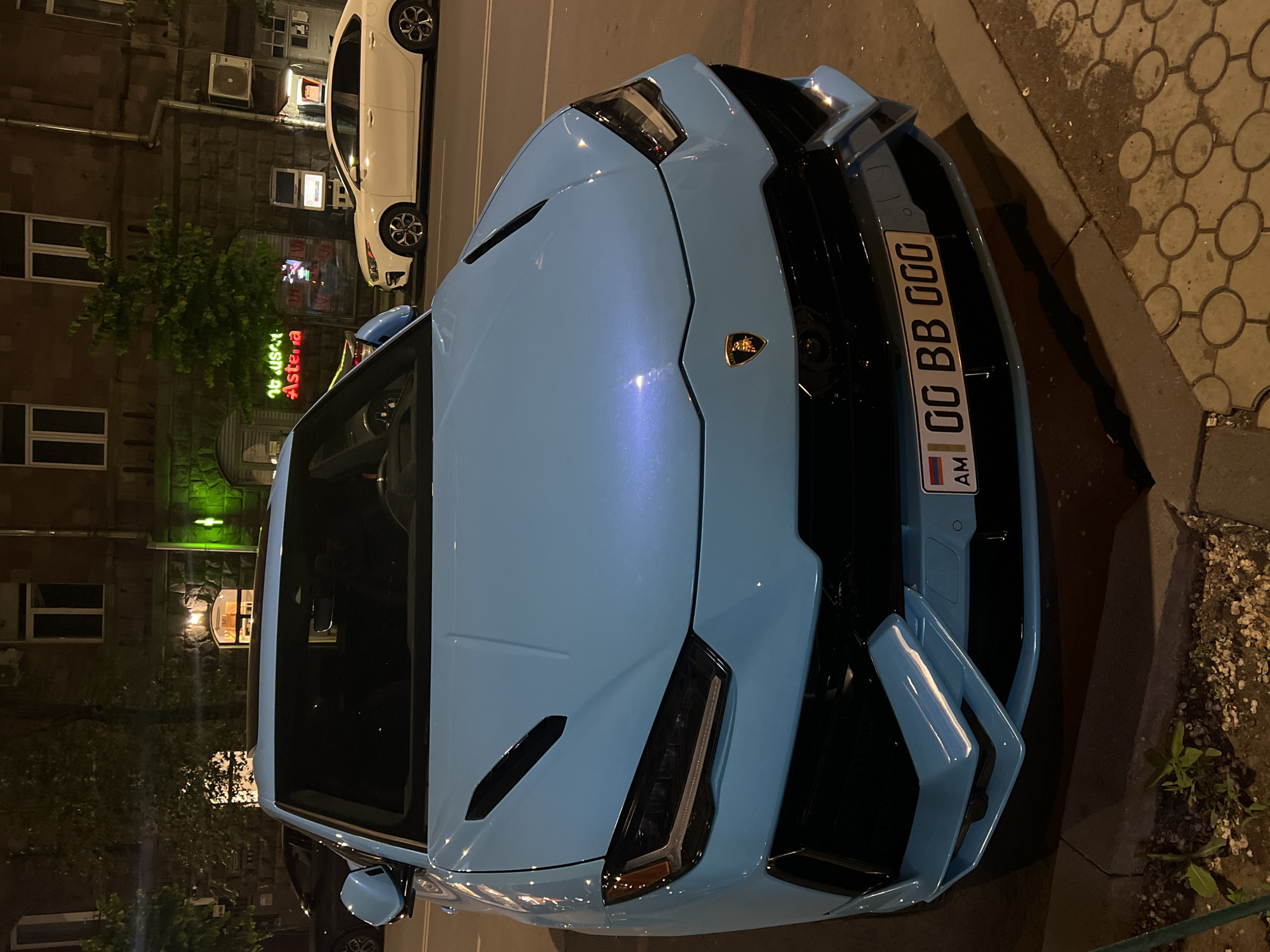 A light blue Lamborghini Urus is parked on a city street at night, in Yerevan, Armenia. The car is positioned front-on, facing the camera. The vehicle has a black grille and black accents on the hood. The license plate reads "00 BB 000" and has an Armenian flag on the left side. Behind the car, a multi-story building with stone facade is visible, with lit windows. A neon sign with the word "Astera" in red is also visible on the building. To the right of the Lamborghini, a white sedan is parked on the street. The street is paved with cobblestones. The overall lighting is dim, suggesting it is nighttime.
