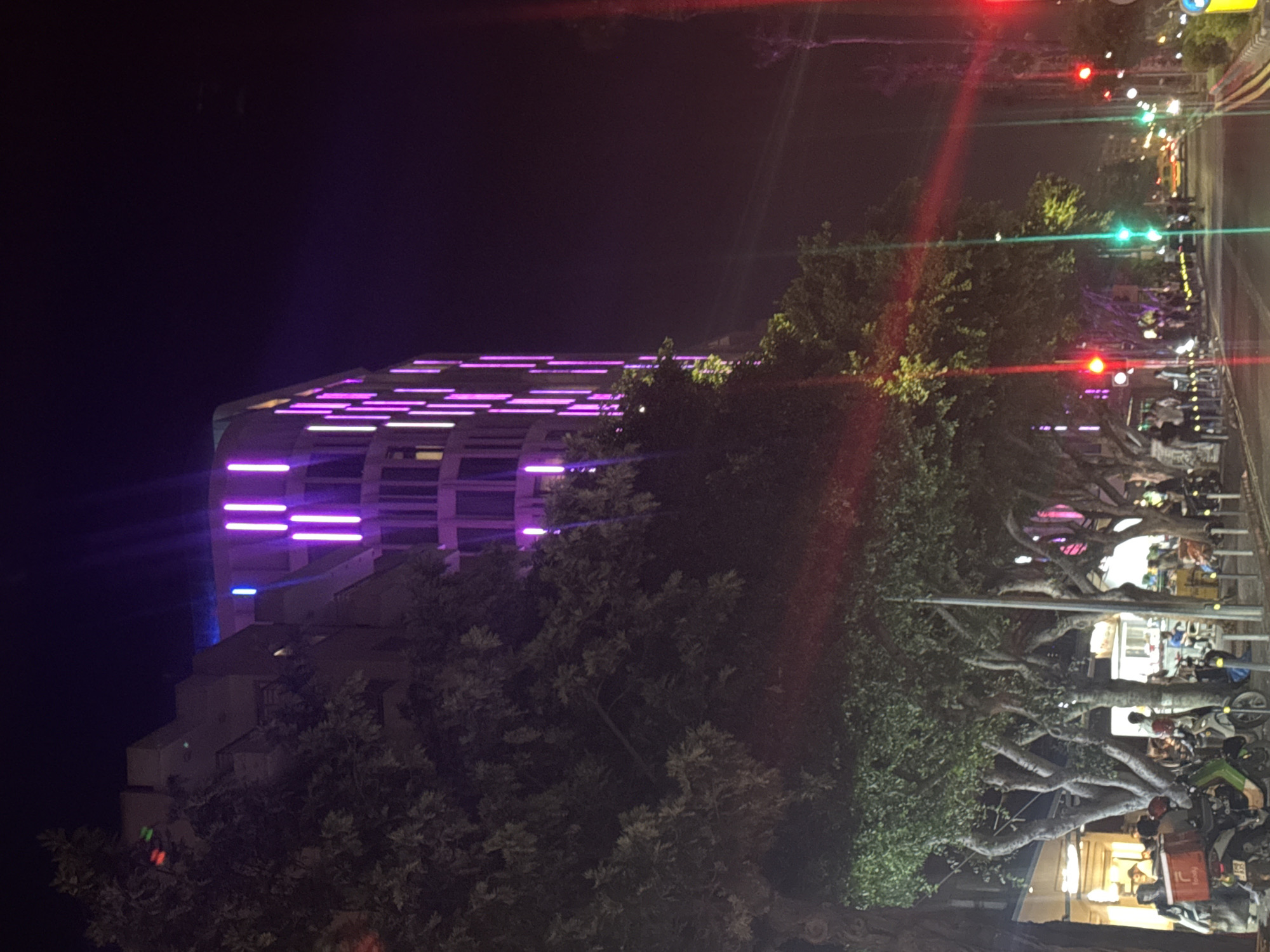 This is a night-time scene on a street in Limassol, Cyprus, likely along the coastal road or a main thoroughfare. A modern building with vertical purple and white lights is prominently featured on the right side of the image. The street is lined with trees that cast shadows, and several motorcycles are parked along the sidewalk. People are visible walking and gathered outside what appear to be establishments. Traffic lights, including red and green signals, are visible along the street. The overall atmosphere suggests a lively urban environment at night, illuminated by artificial lights.