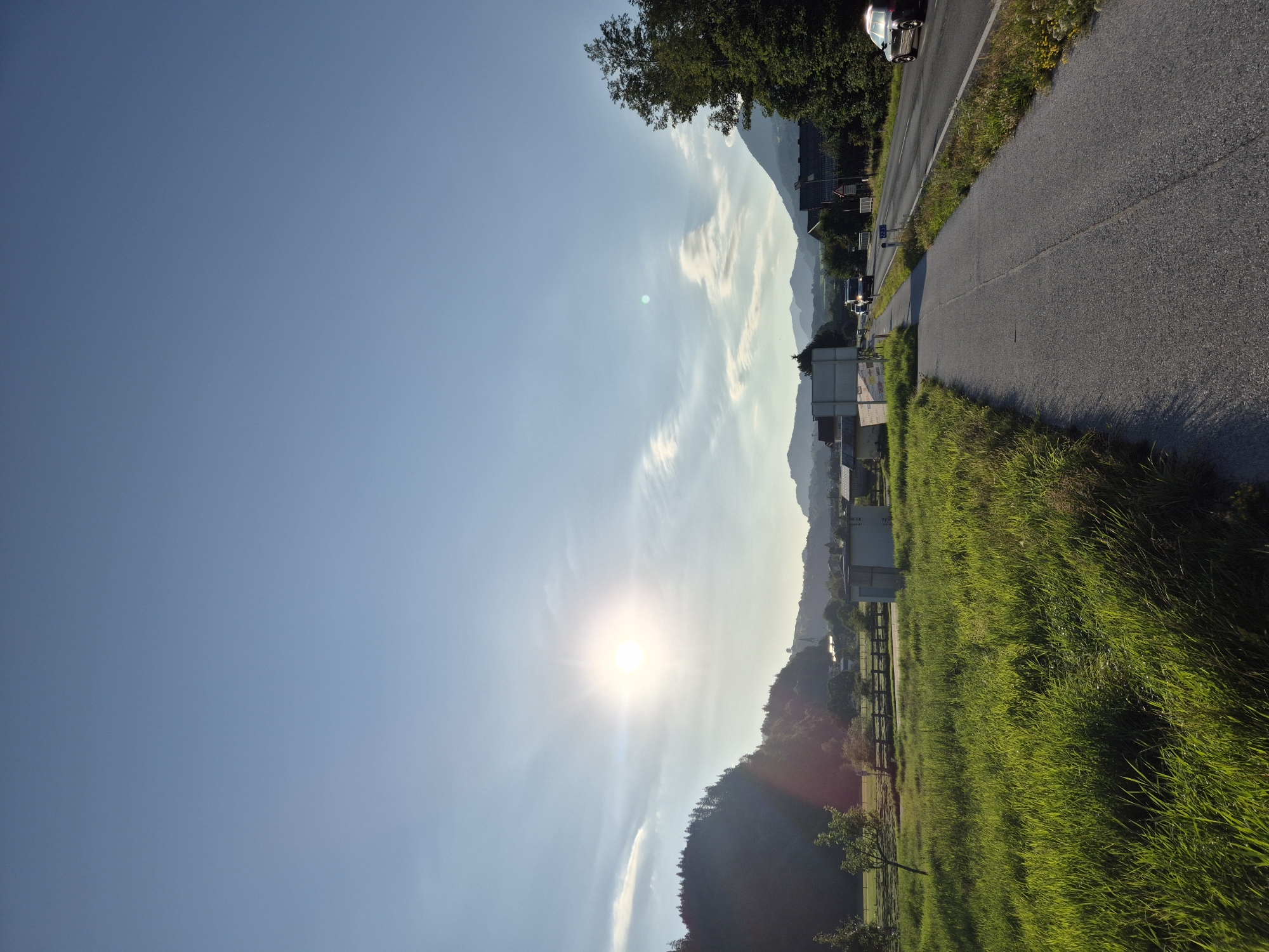 The image depicts a scenic view in Niederndorf, Austria, on a clear day with a bright sun. The foreground features a lush, green grassy field and a paved pathway. To the right, a road runs parallel to the pathway, with a few cars visible. Beyond the road, there are houses and trees, and in the background, rolling hills and mountains are silhouetted against the sky. The sun is low in the sky, casting a warm glow and lens flare across the scene. The overall impression is one of a peaceful rural landscape.
