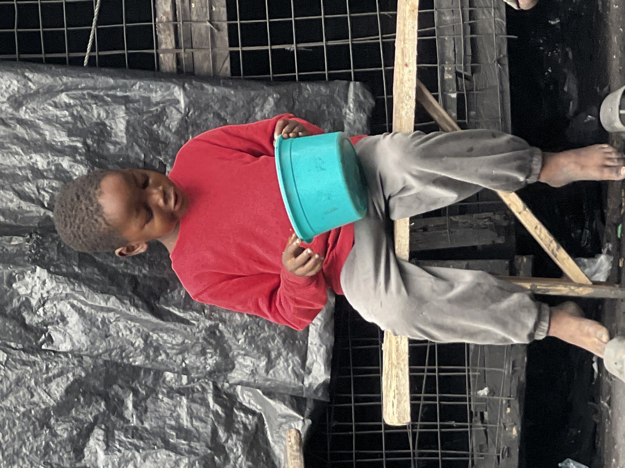 A young boy, appearing to be of African descent and wearing a red long-sleeved shirt and grey sweatpants, sits on a wooden bench. He is holding a teal-colored plastic basin in his lap with both hands. His gaze is directed downwards towards the basin. The bench he is sitting on is positioned in front of a dark, textured background, possibly a tarp or plastic sheeting, with a wire mesh structure visible behind it. The ground below the bench appears to be dirt and littered with debris, including plastic bags and what might be discarded footwear. The scene is set outdoors, possibly in a market or informal settlement area. The overall impression is one of a simple, possibly impoverished, environment.