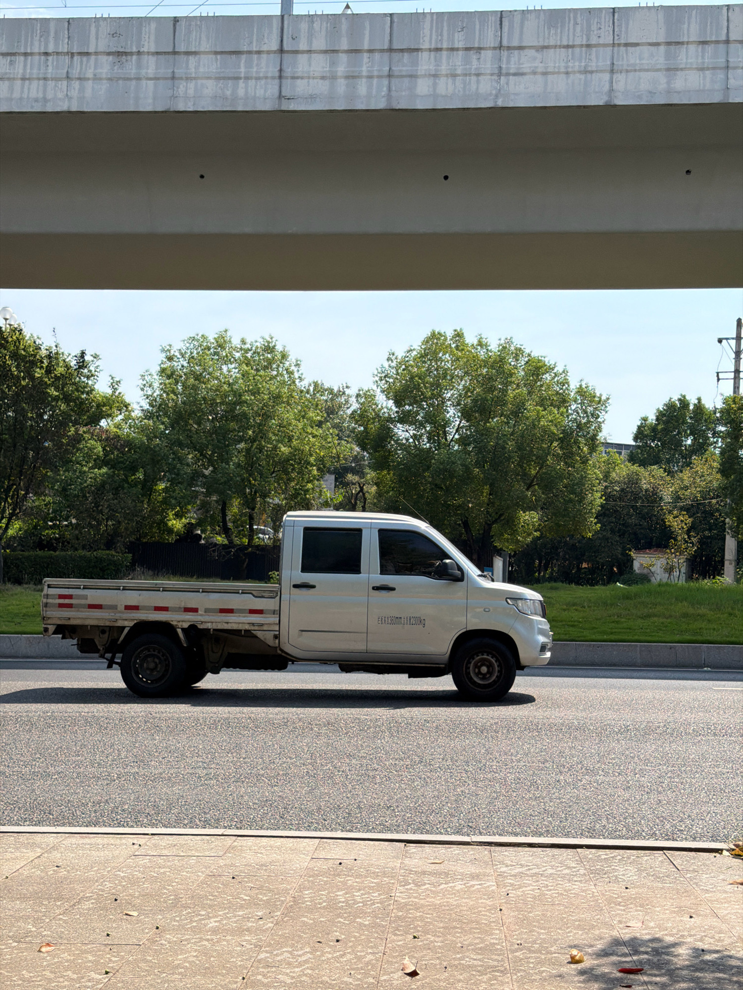 A light-colored pickup truck is captured in profile on a paved road, with a green grassy area and trees in the background. The truck has a single cab with two doors and a long, open bed. The bed of the truck has red and white reflective stripes along its sides. The truck is parked or driving on the right side of the road. Above the scene, a concrete overpass is visible, casting a shadow over the truck and the road. The sky is clear and blue. The location is identified as Jiangjin City, China.