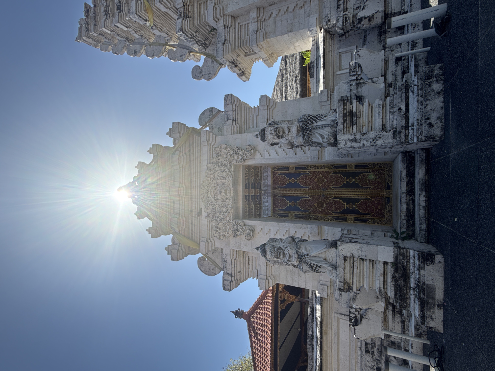 In Kuta Selatan, Indonesia, a sun-drenched, low-angle shot captures the ornate facade of a white stone temple under a clear blue sky. The sun is high and bright, with a lens flare emanating from its center, casting bright rays across the upper portion of the image. The temple entrance is flanked by two stoic stone guardian statues, each wearing traditional checkered cloth around their waists and intricate headdresses. These statues stand on tiered platforms that lead up to the central doorway, which features a large, ornate door with gold and red patterned detailing. Above the doorway, intricate stone carvings adorn the temple's architecture. To the left of the main entrance, a building with a red-tiled, multi-tiered roof is partially visible, suggesting a larger temple complex. The overall scene is one of architectural grandeur and spiritual significance, highlighted by the intense tropical sunlight.