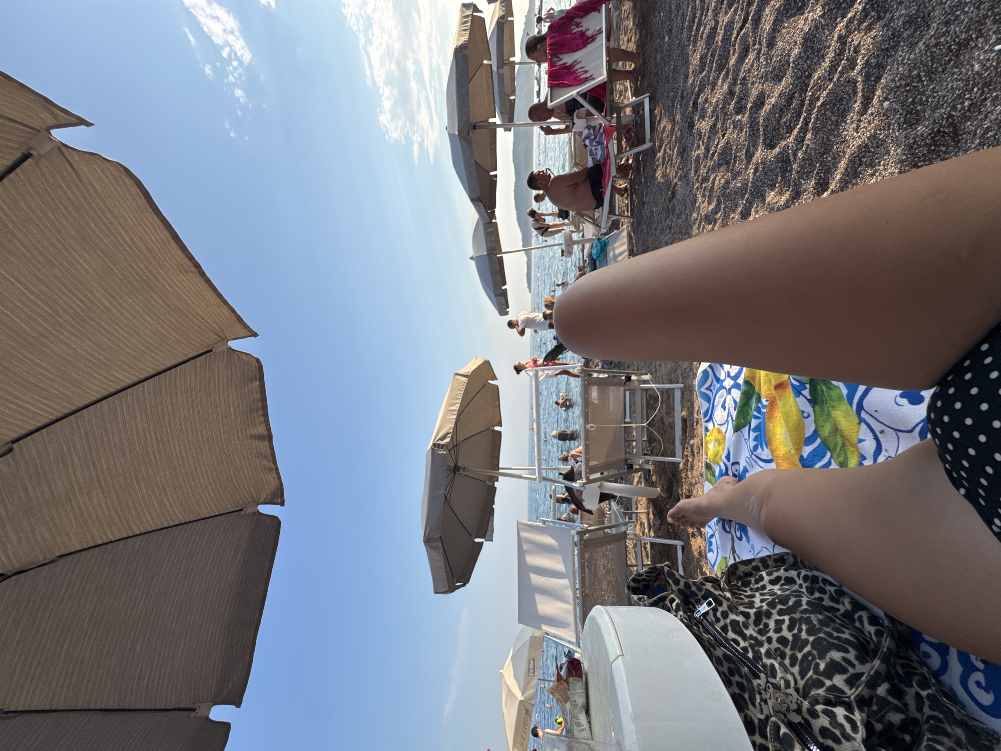 The image is taken from a low-angle, first-person perspective on a beach. A person's legs and feet are in the foreground, lying on a colorful beach towel. To the left, a portion of a white table and a leopard-print bag are visible. Behind the person, a beach scene unfolds with several beach umbrellas, lounge chairs, and people. In the middle ground, the sea stretches out with several people wading or swimming. The sky is blue with a few white clouds, and the sun appears to be low in the sky, casting warm light on the scene. The beach is covered in small pebbles. The location is identified as a beach in Unknown, Unknown.