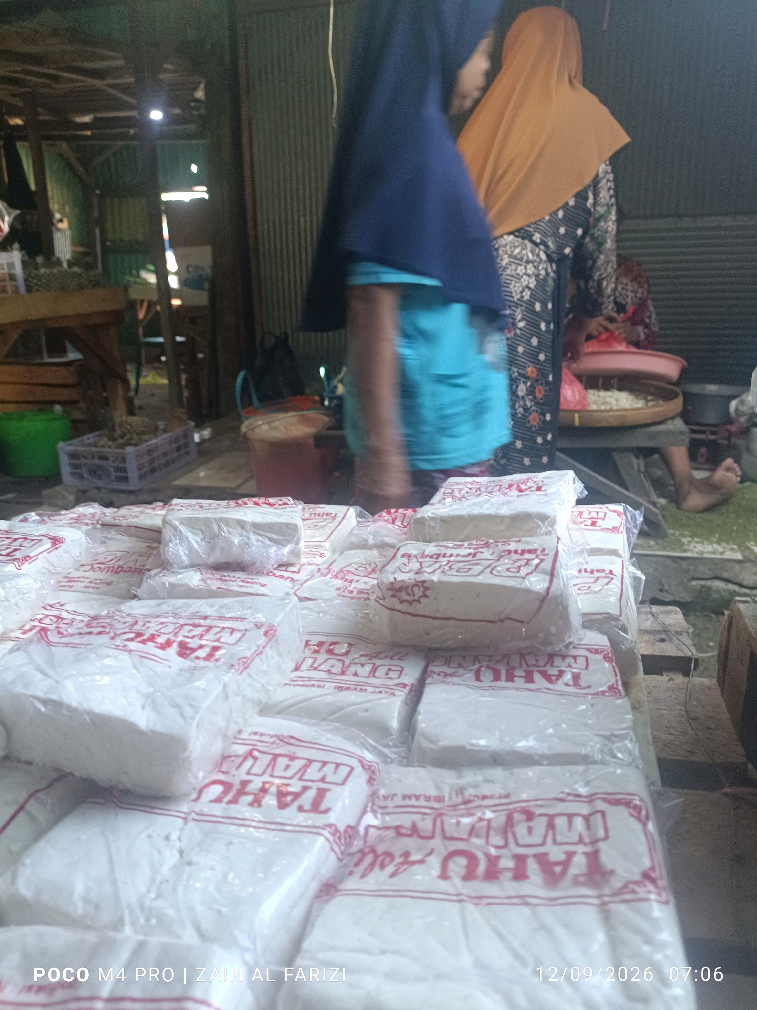 The image depicts an outdoor market stall in Arosbaya, Indonesia, likely a local food vendor. In the foreground, stacked white blocks, identified by the red text "TAHU MALANG" (Malang Tofu) printed on their packaging, are prominently displayed. These appear to be individually wrapped tofu blocks. To the left, a green bucket and a woven plastic crate are visible. In the background, two individuals are partially visible. One is wearing a blue hijab and a light blue shirt. The other is wearing an orange hijab and a patterned dress. The background also reveals a corrugated metal wall and wooden structures, suggesting a rustic or traditional market setting. A woman's bare foot is visible towards the right side of the frame, suggesting she is a vendor or customer. The lighting suggests it is daytime. The timestamp "12/09/2026 07:06" indicates the image was taken on September 12, 2026, at 7:06 AM. The text "POCO M4 PRO | ZAIN AL FARIZI" likely refers to the phone used to capture the image and the photographer's name.