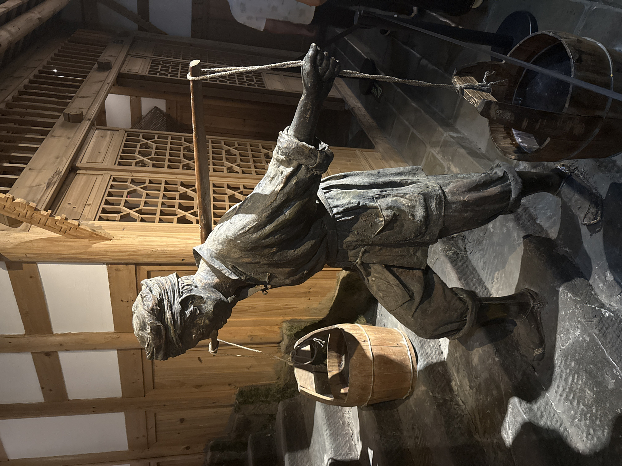 The image depicts a statue of a person carrying two wooden buckets by a shoulder yoke, a common method of transporting water in historical China. The statue is positioned in what appears to be a museum or heritage site, with traditional wooden architecture visible in the background, characterized by latticed screens and carved wooden beams. Stone steps are visible to the left, suggesting an outdoor or semi-outdoor setting. The statue is made of a dark material, possibly bronze or aged wood, and is realistically detailed, showing folds in the clothing and the texture of the yoke and buckets. The buckets are made of wood with metal reinforcements and rope handles. The lighting suggests an indoor environment, possibly with artificial light highlighting the statue and its surroundings. While the specific location within Yuzhong District, China, is not explicitly identifiable from the image alone, the architectural style points to a historical or cultural exhibition space.