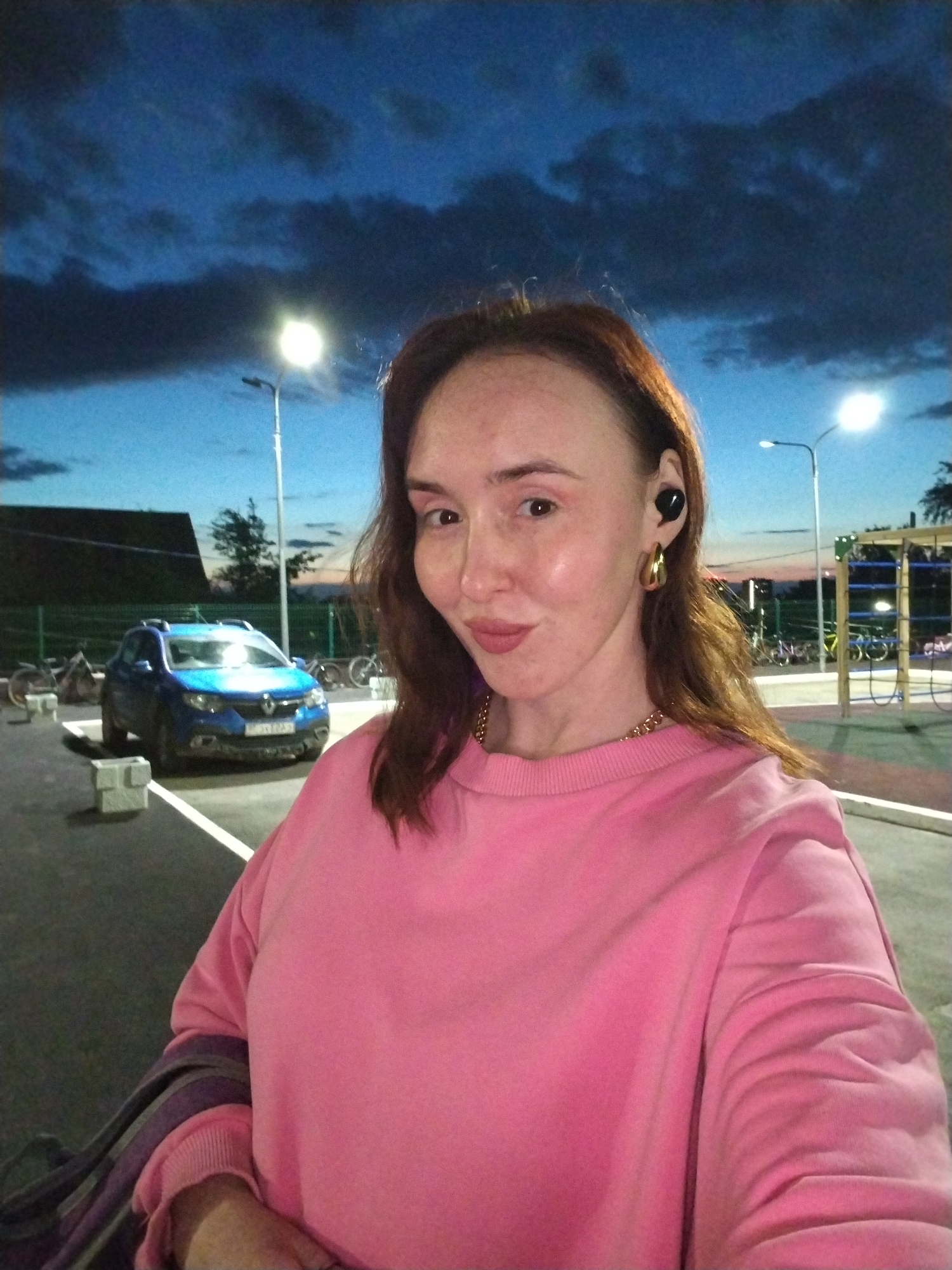 The image is a selfie of a woman taken outdoors at dusk or night. The woman has reddish-brown hair and is wearing a bright pink long-sleeved sweatshirt. She accessorizes with a gold-colored chain necklace, a black earbud in her left ear, and a gold-colored hoop earring in her right ear. She looks towards the camera with a faint smile.
The background features a dark blue sky with visible dark clouds, gradually lightening towards the horizon. Multiple bright streetlights illuminate the scene. To the left, a blue Renault Duster SUV is parked, with its front lights reflecting light. Concrete blocks are visible on the ground near the car. Further back, a green mesh fence runs across the mid-ground, behind which several bicycles are parked. On the right side of the background, metal playground or fitness equipment can be seen. The ground is a paved surface, likely asphalt, with white painted lines indicating parking spaces. The license plate on the blue car has visible Cyrillic characters but is not legible. The scene depicts an outdoor area, possibly a community space or parking lot adjacent to a recreational area.